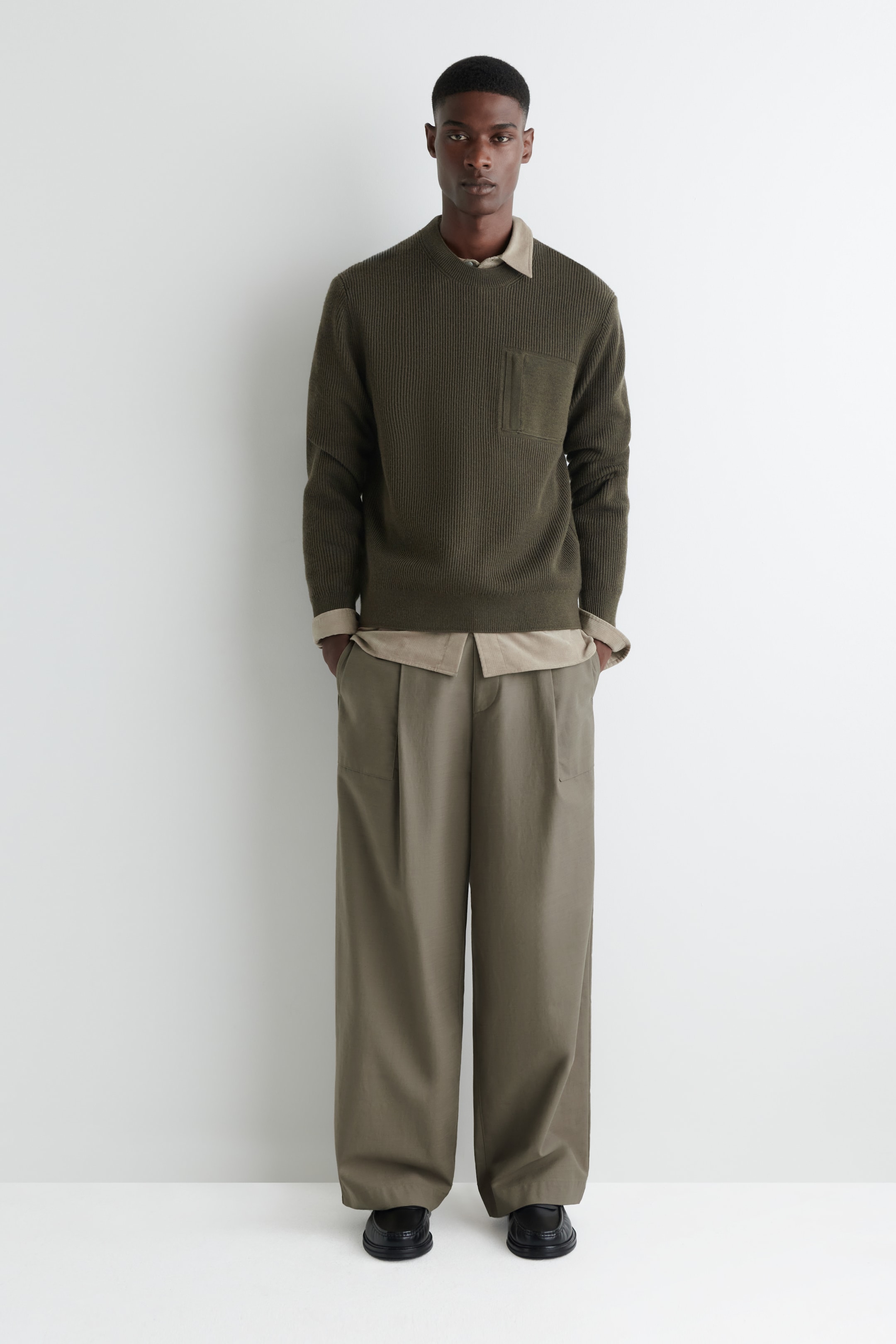 Grösseres Bild ansehen: Ein olivgrüner Pullover wird aus einem frontalen Winkel präsentiert, mit einem vertikalen Rippstrick, einem Rundhalsausschnitt und einer aufgesetzten Tasche auf der linken Brust. Er hat gerippte Bündchen und einen gerippten Saum und wird über einem hellen Hemd mit sichtbarem Kragen und sichtbaren Manschetten getragen.