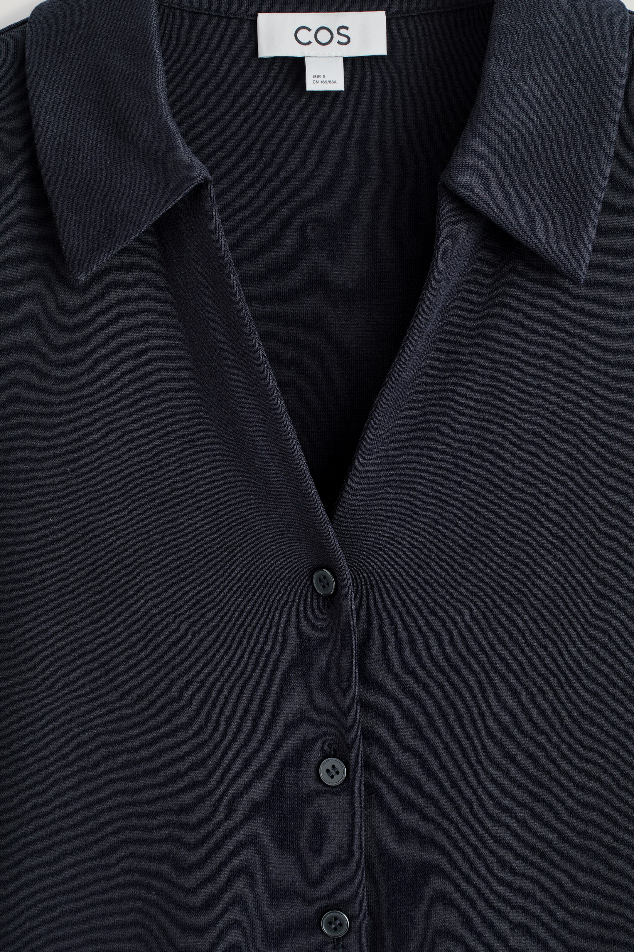 Agrandir l'image: Une chemise bleu foncé, confectionnée dans un tissu tricoté lisse, est présentée de face. Elle présente un col ouvert formant un décolleté en V et une patte de boutonnage à l'avant avec trois boutons ton sur ton visibles. Une étiquette de marque blanche est également visible à l'arrière du cou.