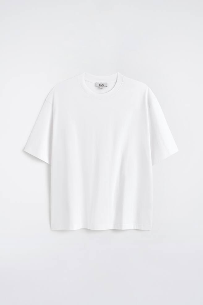 Een wit t-shirt wordt frontaal getoond, met een geribbelde ronde hals, korte mouwen met verlaagde schoudernaden en een ontspannen pasvorm, met een klein 'COS' label zichtbaar aan de achterkant van de hals. - 1