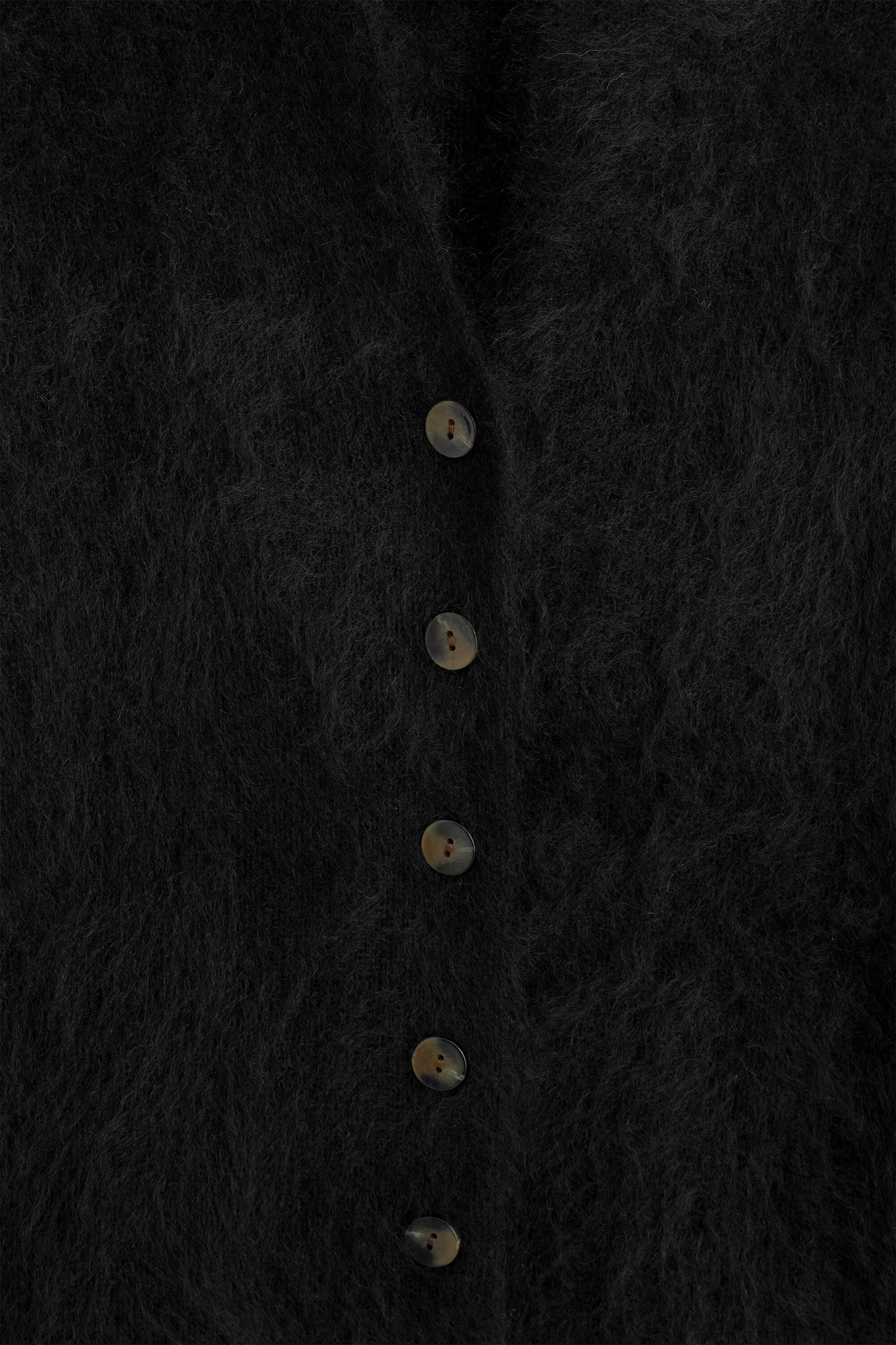 The draped brushed merino wool cardigan is shown in a close-up front view, highlighting its soft, fuzzy black texture. A vertical row of five dark, marbled buttons runs down the front closure of the garment. The draped brushed merino wool cardigan is shown in a close-up front view, highlighting its soft, fuzzy black texture. A vertical row of five dark, marbled buttons runs down the front closure of the garment.