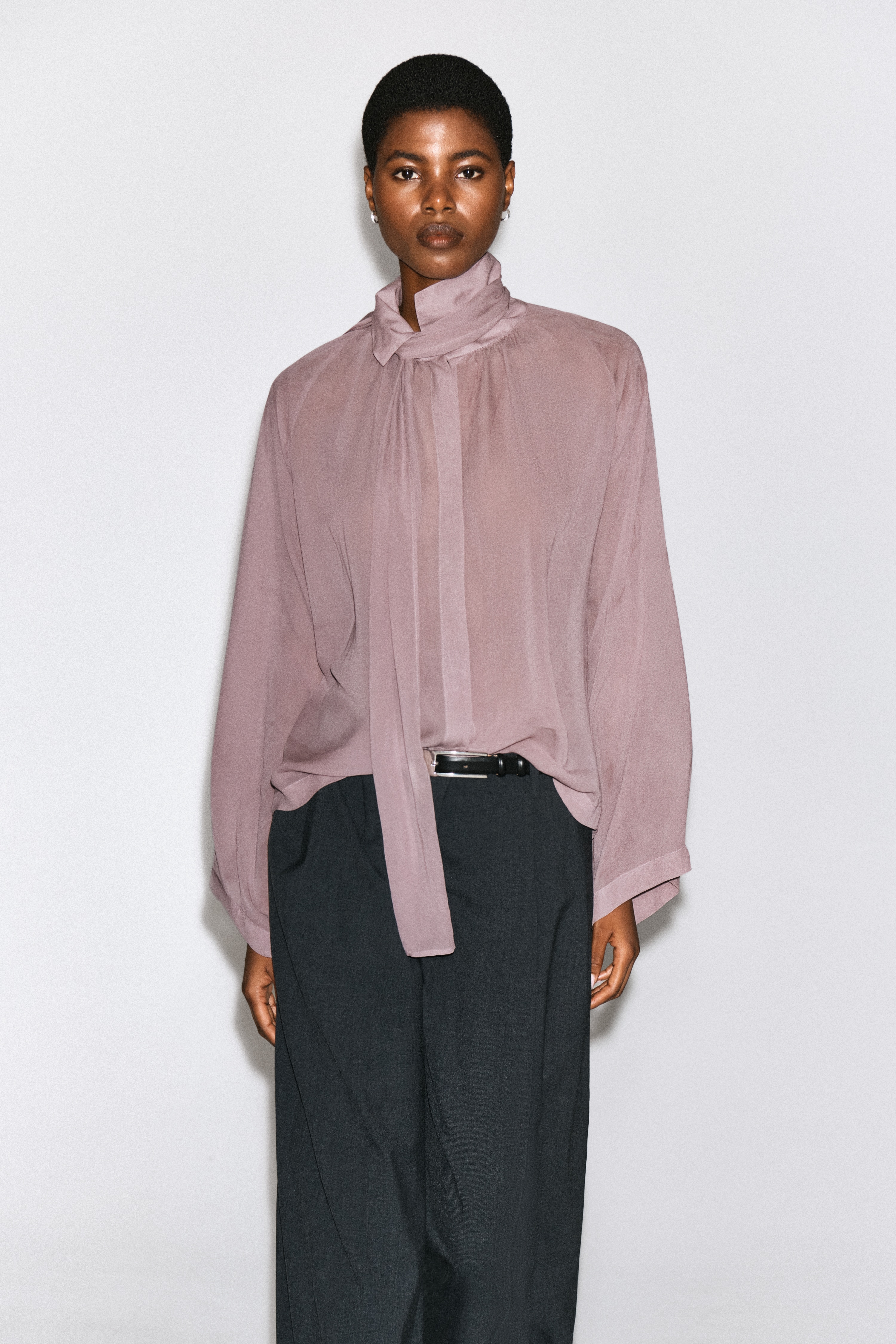 Le haut fluide Ã  dÃ©tail foulard est prÃ©sentÃ© de face dans une teinte mauve poussiÃ©reuse, avec un col pointu et deux longs pans assortis en tissu qui retombent sur la poitrine. La blouse affiche une silhouette dÃ©contractÃ©e, avec des manches larges et fluides ainsi quâune patte de boutonnage frontale dissimulÃ©e.