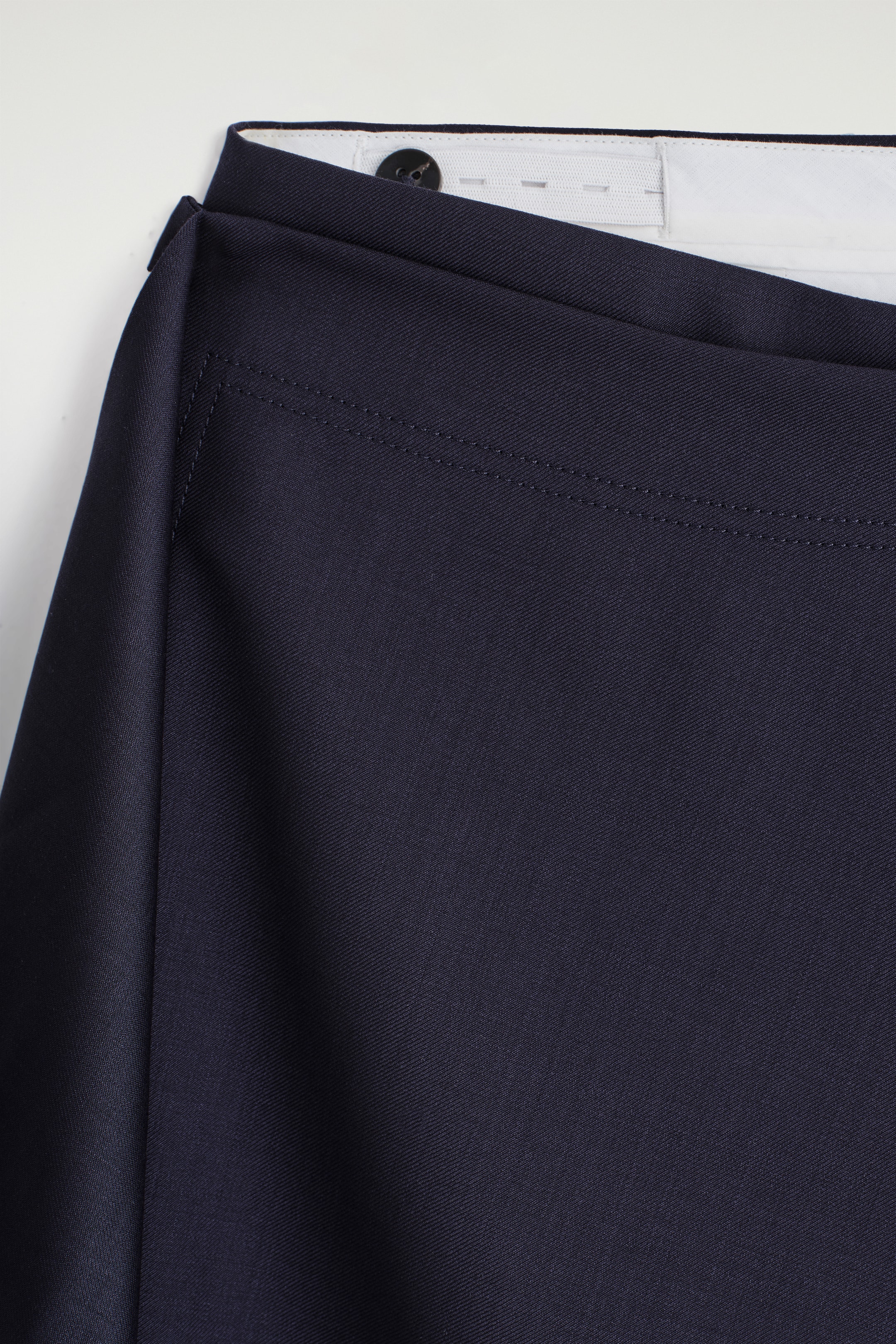 View larger image: A close-up view of the front of dark blue trousers is showing a draped skirt detail wrapping around the waist. A black button is visible on the inner waistband, and two parallel stitch lines are running horizontally across the fabric below the waist.