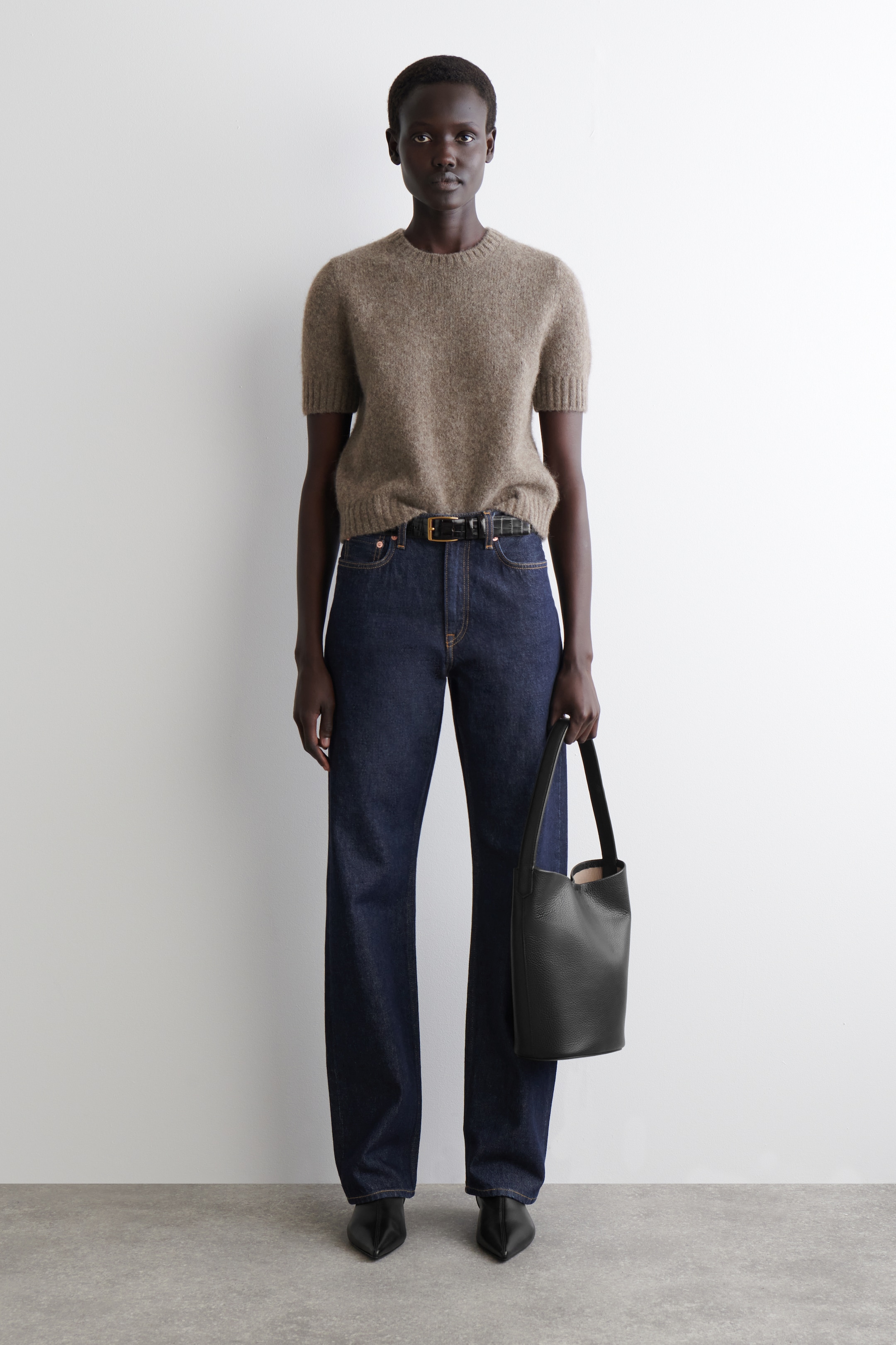 Agrandir l'image: Une tenue complète, vue de face, est présentée, mettant en avant un haut en tricot duveteux à manches courtes de couleur brun clair, avec un col rond et une bordure côtelée. Ce haut est associé à un jean à coupe droite et lavage foncé, une ceinture texturée noire avec une boucle dorée, et des bottes noires à bout pointu. Un sac seau noir en cuir grainé est tenu.