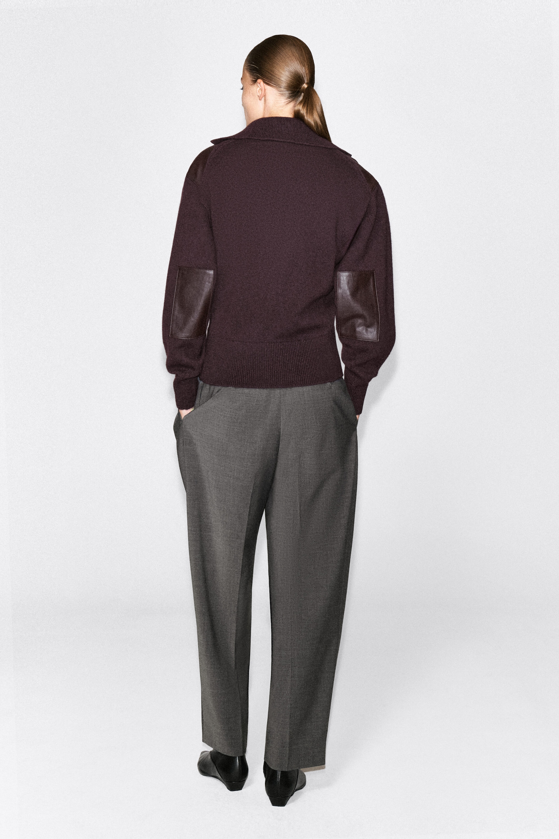 A dark burgundy merino wool zip-up sweater is being worn by the model, featuring dark brown leather patches on the elbows and a ribbed hem as seen from the back. The sweater is paired with gray wide-leg trousers and black shoes. A dark burgundy merino wool zip-up sweater is being worn by the model, featuring dark brown leather patches on the elbows and a ribbed hem as seen from the back. The sweater is paired with gray wide-leg trousers and black shoes.
