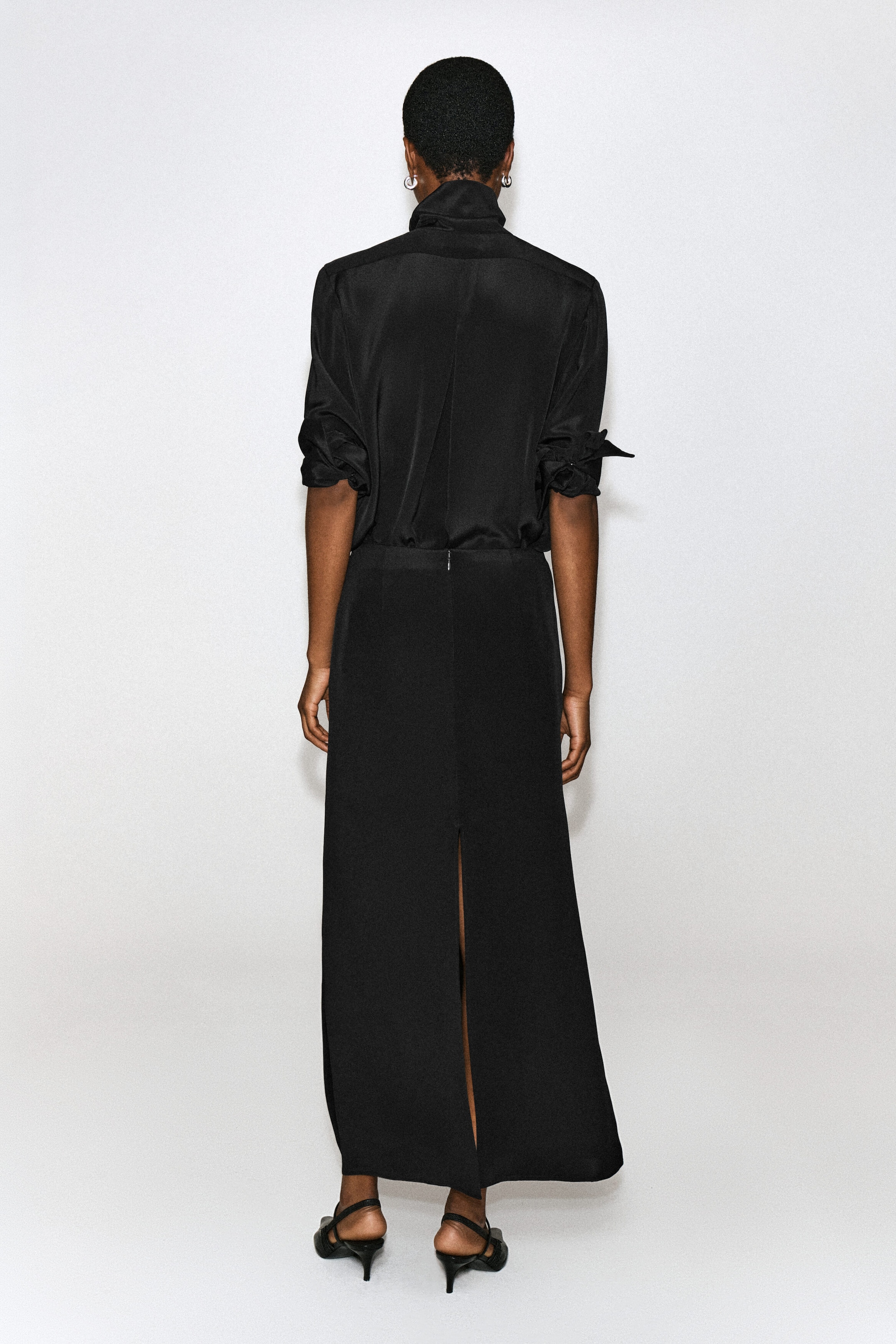 A black maxi skirt with a straight cut and a high centre-back slit is worn by the model, paired with a tucked-in black long-sleeve blouse with rolled-up sleeves. The skirt features a small back zip at the waist, and the look is completed with black heeled slingback sandals.