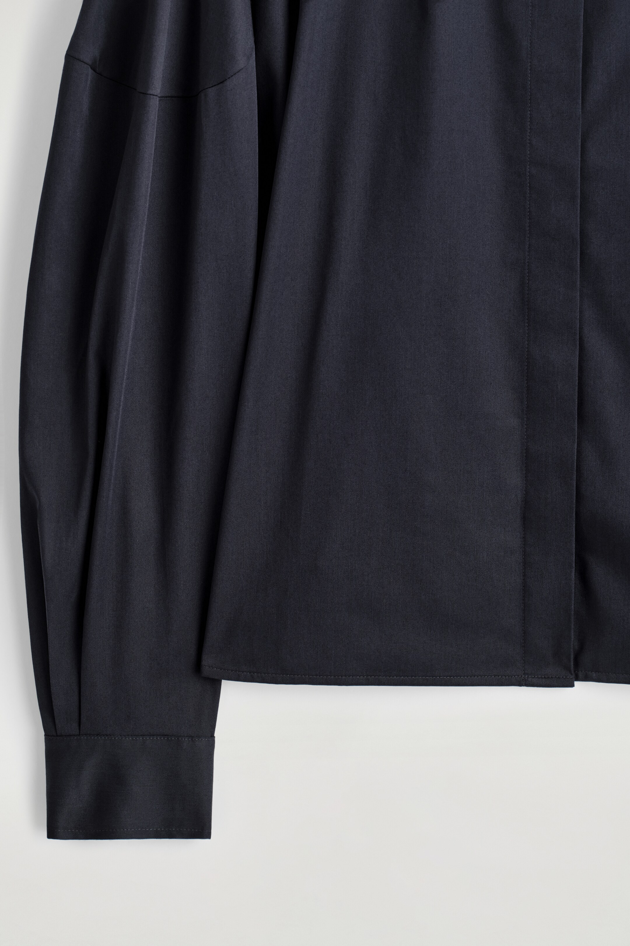 Agrandir l'image: Une chemise noire est dotée d'une manche raglan volumineuse qui se termine par un poignet large et structuré. Le corps de la chemise présente un détail subtil de pli vertical sur le devant et un ourlet droit.