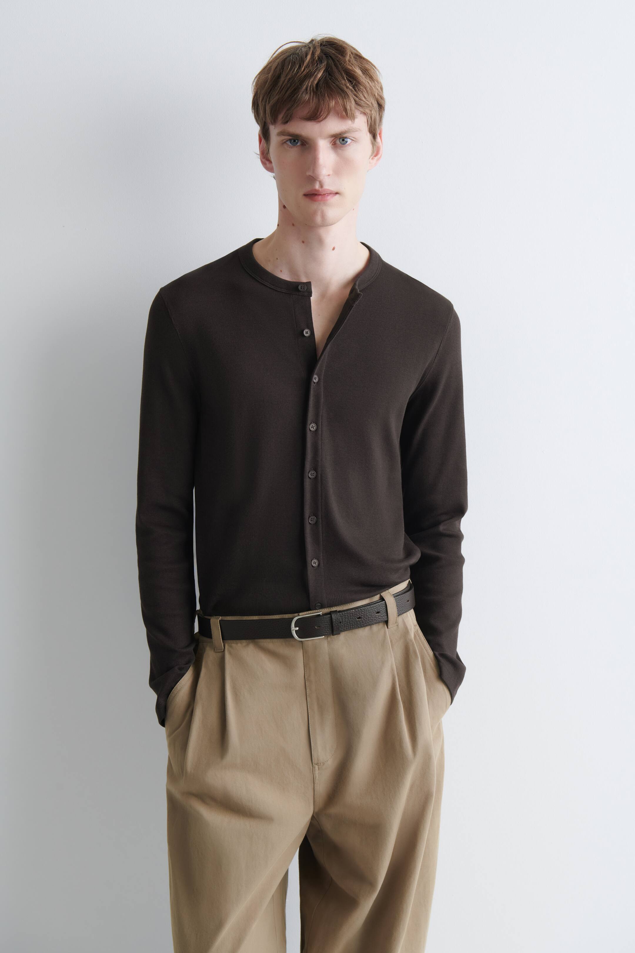 Agrandir l'image: Un cardigan marron foncé, ajusté et finement côtelé, avec un col sans col et une fermeture boutonnée sur le devant, est porté par le mannequin. Plusieurs boutons du haut sont déboutonnés, et le cardigan est rentré dans un pantalon plissé kaki.