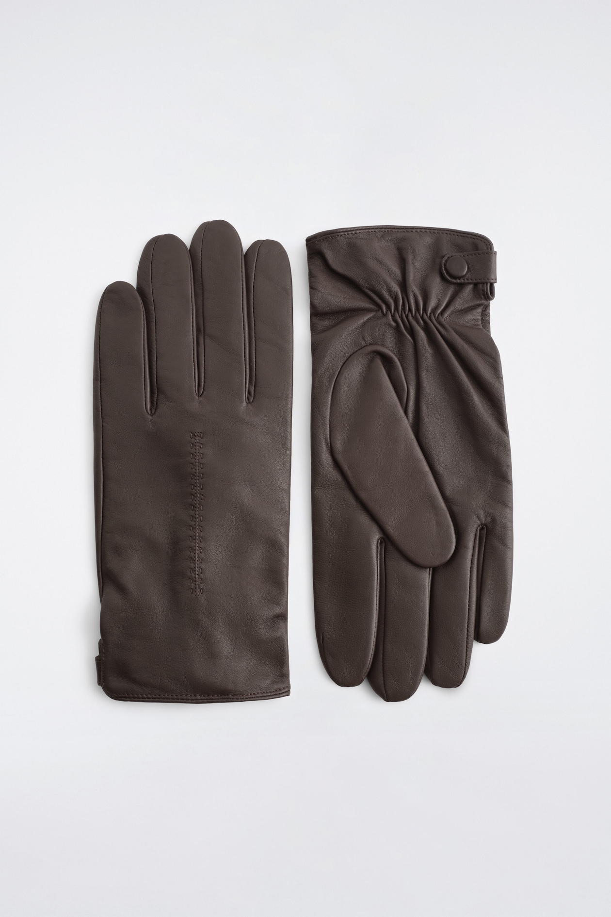 A pair of dark brown leather gloves is laid flat, with one glove showing the back of the hand with subtle vertical topstitching, and the other showing the palm side with a gathered wrist and a leather-covered snap closure.