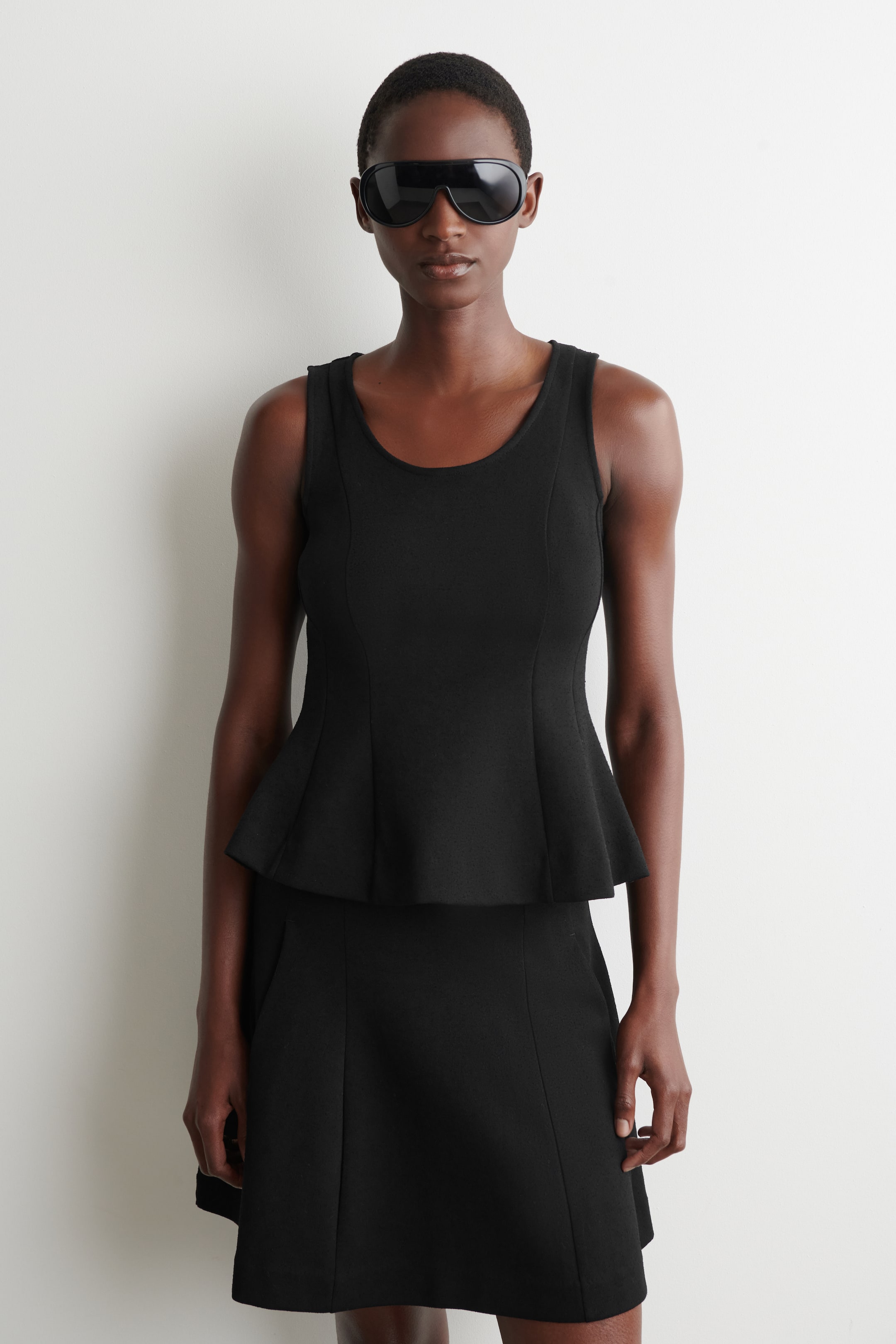 Ver imagen más grande: Un top negro sin mangas de estilo peplum presenta un escote redondo y un cuerpo ajustado con paneles, que se ensancha en un dobladillo peplum estructurado. Se combina con una minifalda negra a juego.