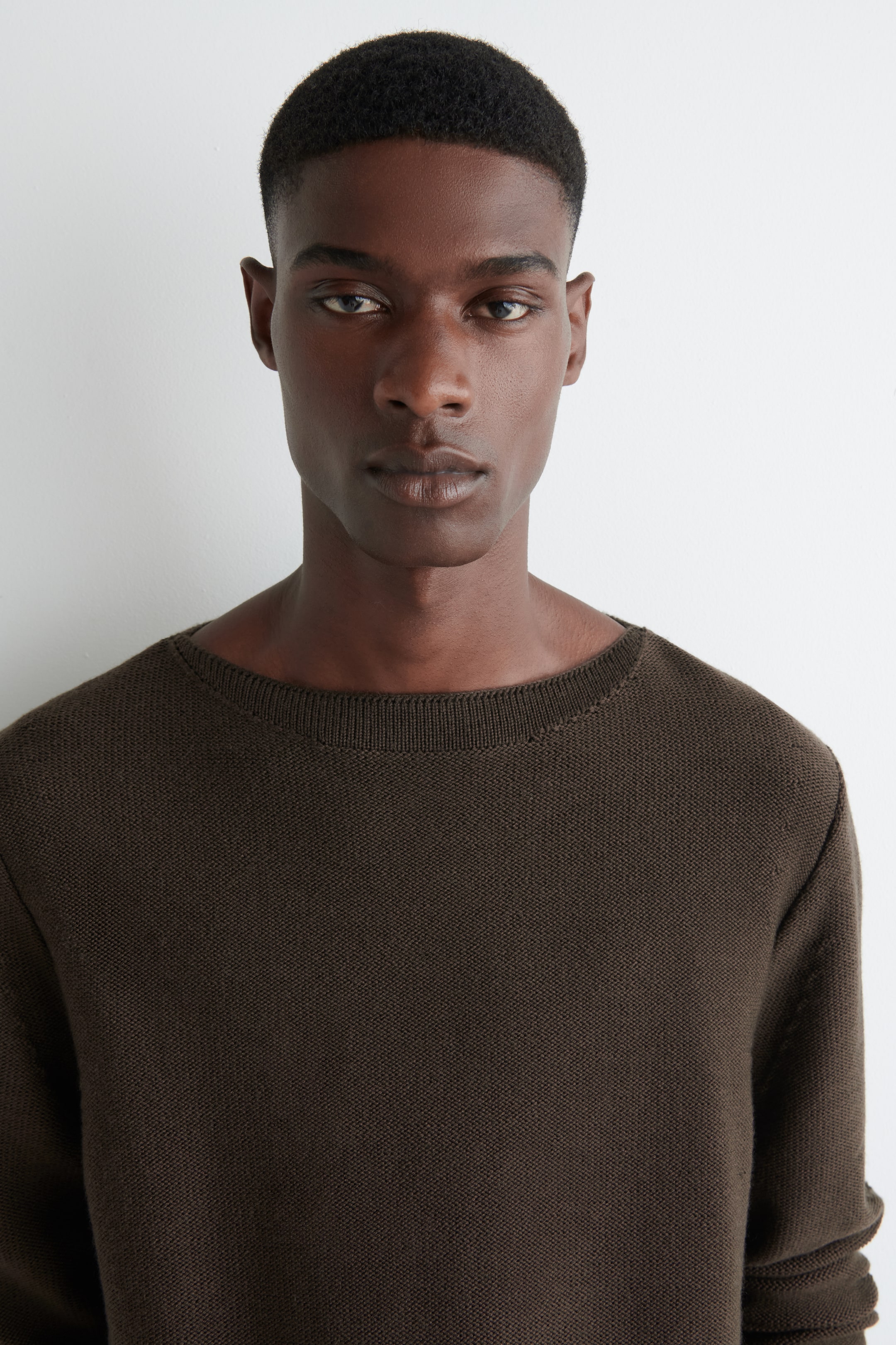 Grösseres Bild ansehen: Ein dunkelbrauner Strickpullover mit Textur wird getragen, nach vorne gerichtet, mit einem breiten, gerippten Rundhalsausschnitt.