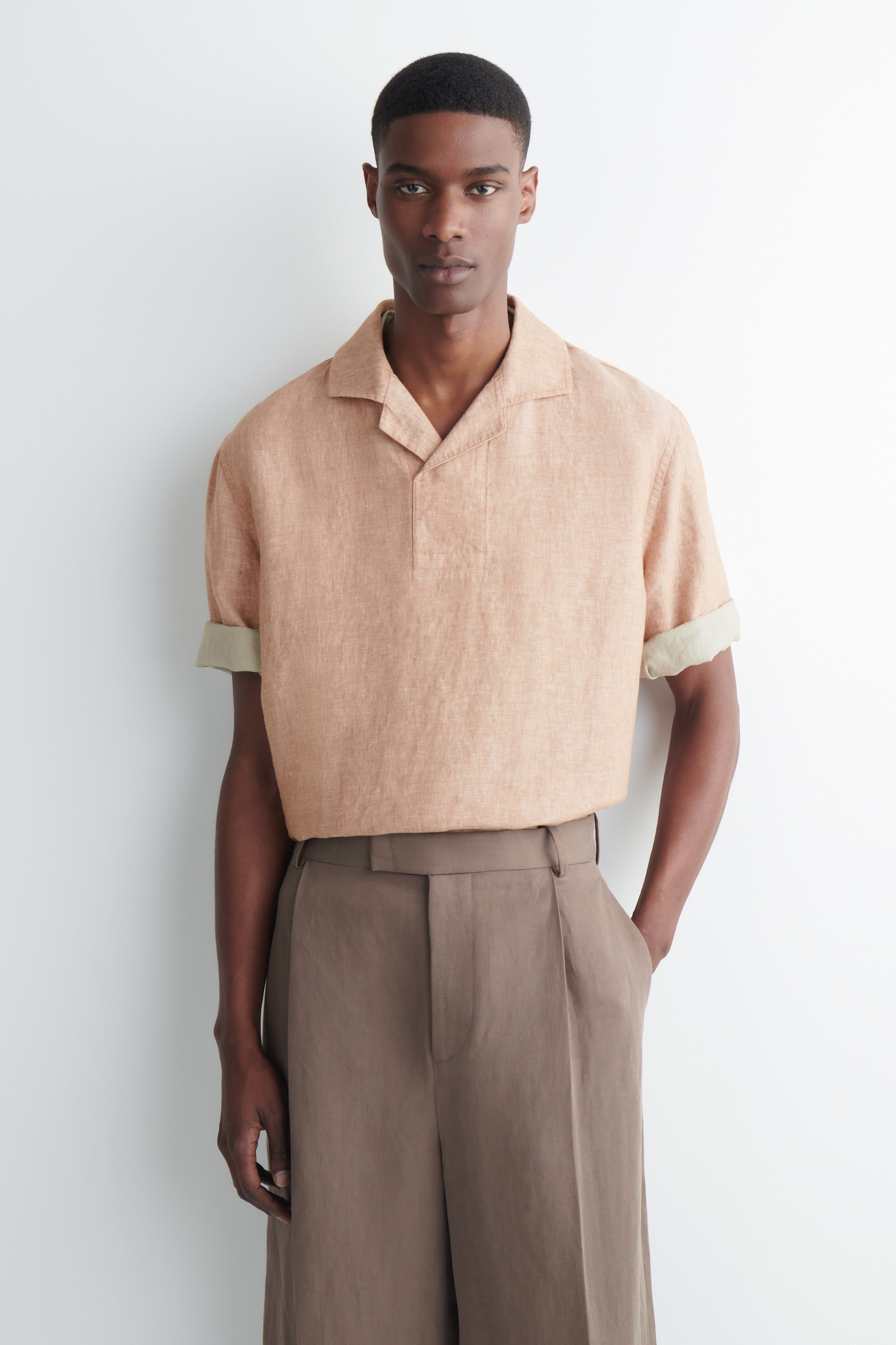 Agrandir l'image: Une chemise marron clair texturée présente un col à revers, une patte partielle et des manches courtes retroussées révélant un tissu intérieur plus clair. Elle est rentrée dans un pantalon plissé marron à jambes larges avec des passants de ceinture, les deux vêtements faisant face vers l'avant.