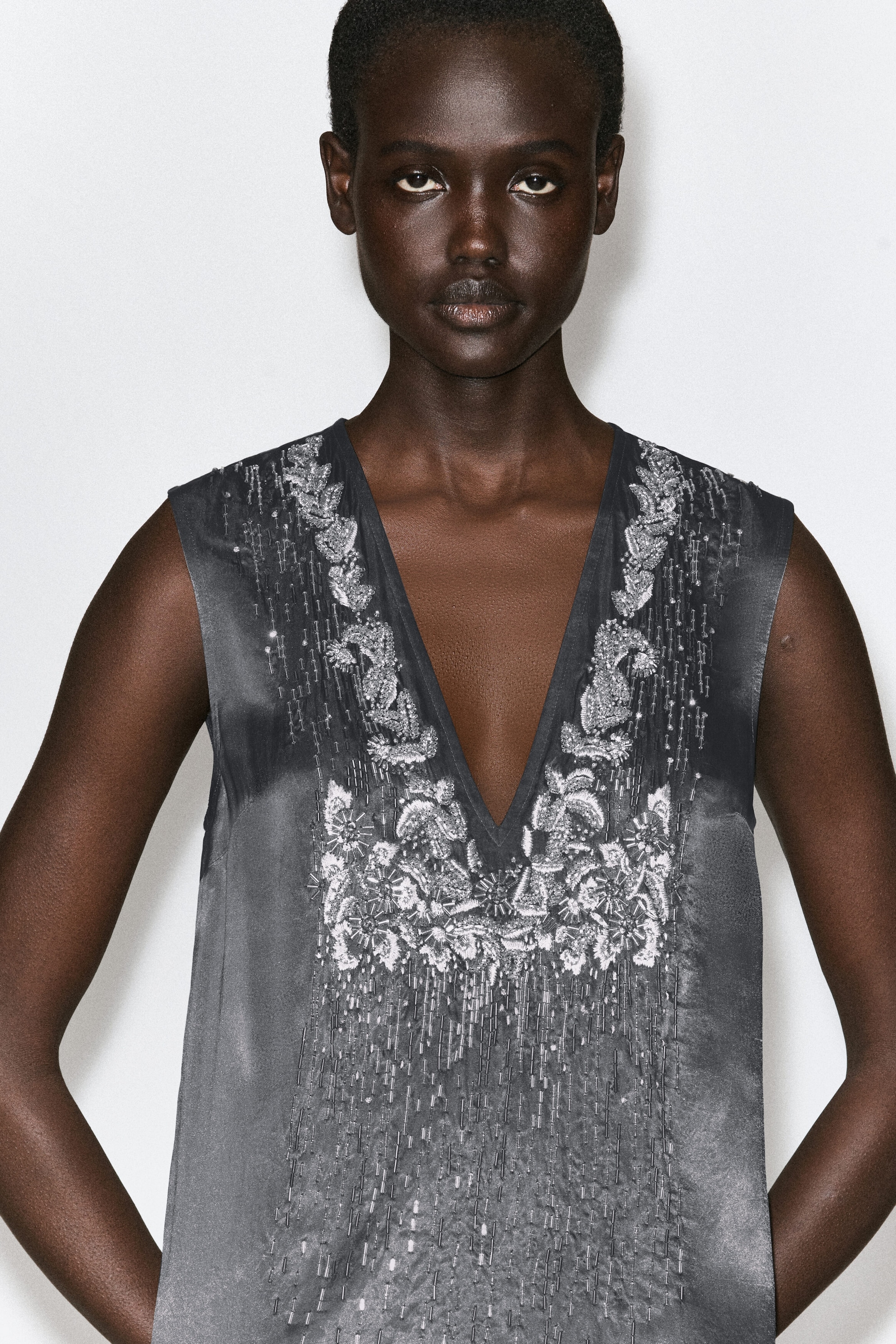 A dark gray beaded satin V-neck midi dress is shown from the front, featuring intricate silver floral embroidery and beadwork framing a deep V-neckline. Vertical rows of silver beads cascade down the front of the sleeveless bodice. A dark gray beaded satin V-neck midi dress is shown from the front, featuring intricate silver floral embroidery and beadwork framing a deep V-neckline. Vertical rows of silver beads cascade down the front of the sleeveless bodice.