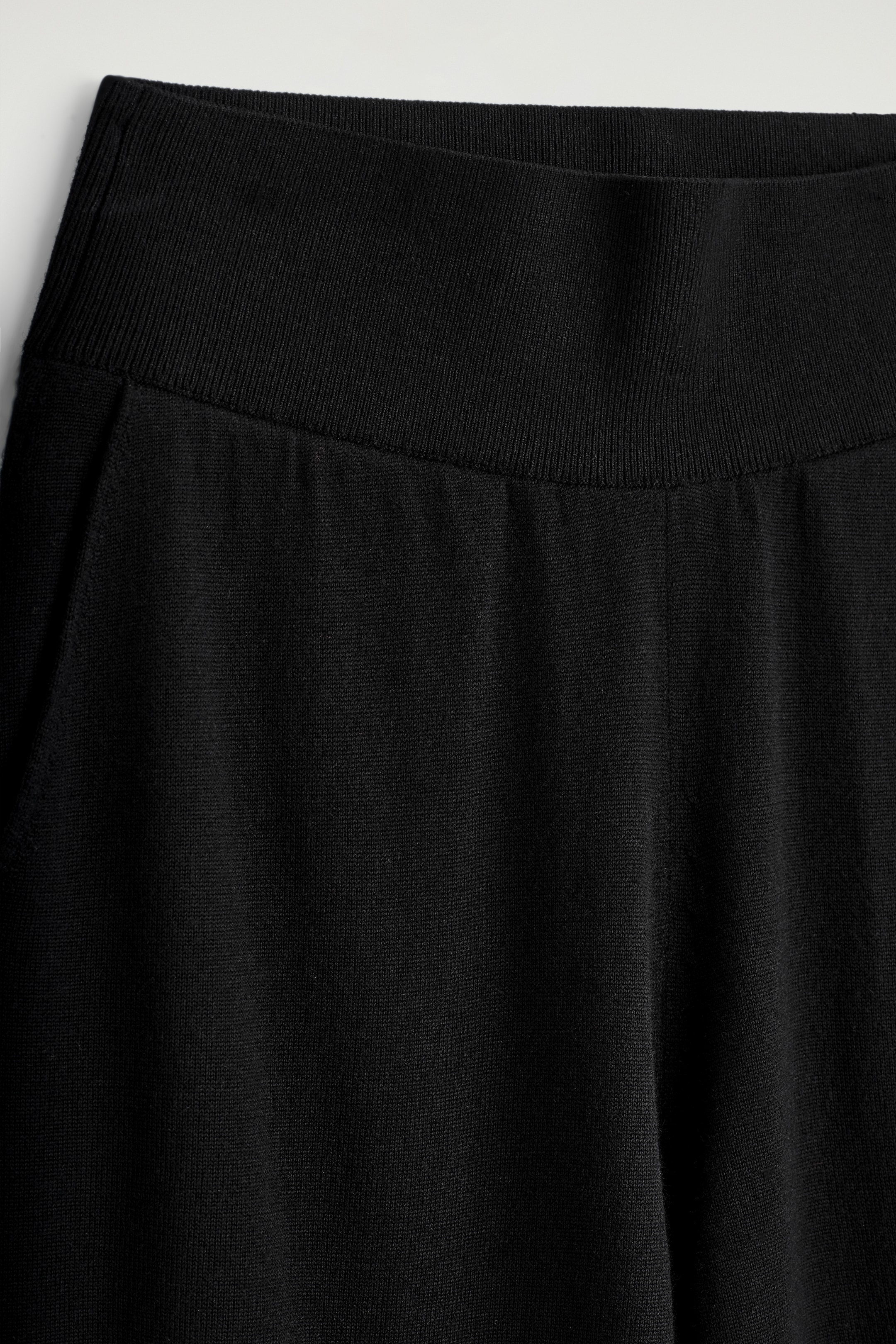 Visualizza immagine più grande: I pantaloni in lana merino nera sono mostrati dalla parte anteriore, con una fascia in vita ampia e a coste. Una tasca laterale è visibile sul lato destro del capo, e la fine trama lavorata a maglia del tessuto è evidente.