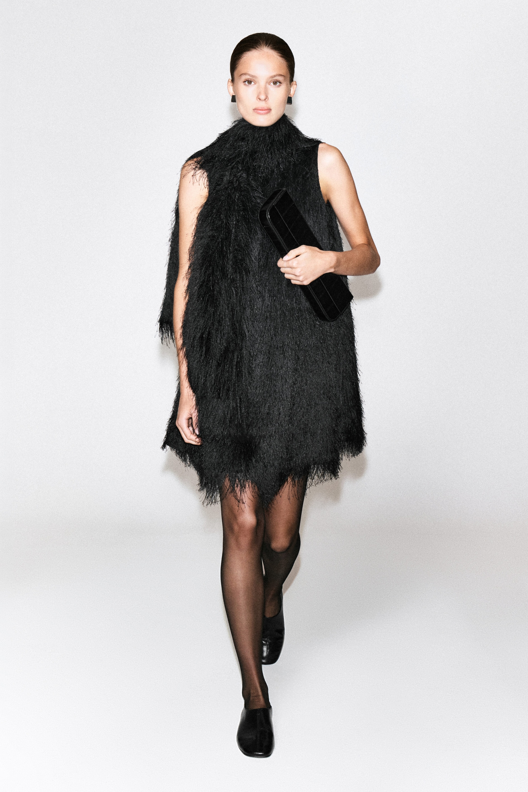 Ein schwarzes Mini-Bodycon-Kleid mit Fransen, mit hohem, strukturiertem Kragen und ärmelloser Silhouette, bedeckt mit langen, feinen Fransen, wird von dem Model getragen. Das Model geht nach vorne und hält eine schwarze Clutch in der Hand. Dazu trägt es transparente schwarze Strumpfhosen und schwarze flache Schuhe. Ein schwarzes Mini-Bodycon-Kleid mit Fransen, mit hohem, strukturiertem Kragen und ärmelloser Silhouette, bedeckt mit langen, feinen Fransen, wird von dem Model getragen. Das Model geht nach vorne und hält eine schwarze Clutch in der Hand. Dazu trägt es transparente schwarze Strumpfhosen und schwarze flache Schuhe.