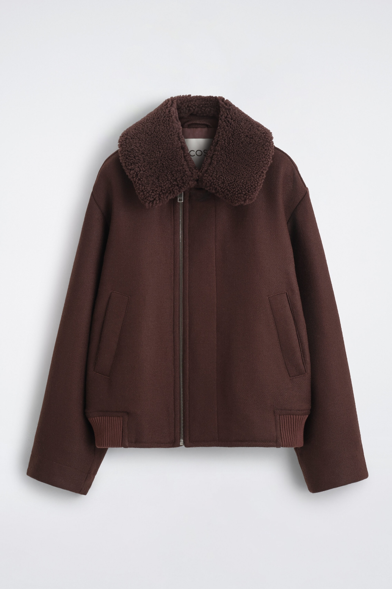 A dark brown bomber jacket is shown from the front, featuring a plush, textured shearling-style collar and an asymmetrical front zipper. The jacket features dropped shoulders, wide sleeves, diagonal welt pockets, and a ribbed hem detail.