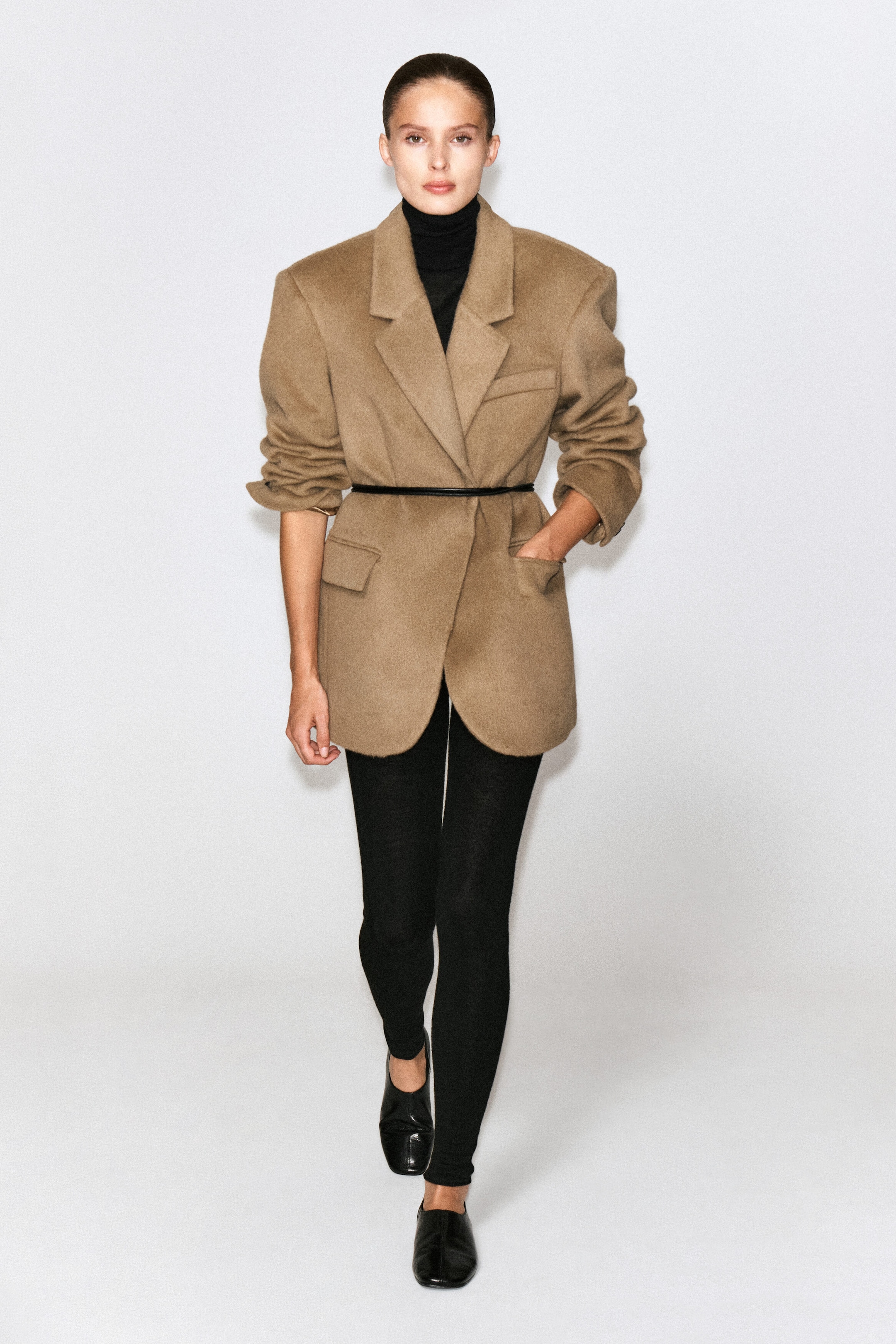 A camel-colored brushed wool blazer featuring an oversized silhouette, notched lapels, and structured shoulders is worn by the model. The blazer is cinched at the waist with a thin black belt and layered over a black turtleneck and leggings, with the model placing one hand in a side flap pocket. A camel-colored brushed wool blazer featuring an oversized silhouette, notched lapels, and structured shoulders is worn by the model. The blazer is cinched at the waist with a thin black belt and layered over a black turtleneck and leggings, with the model placing one hand in a side flap pocket.