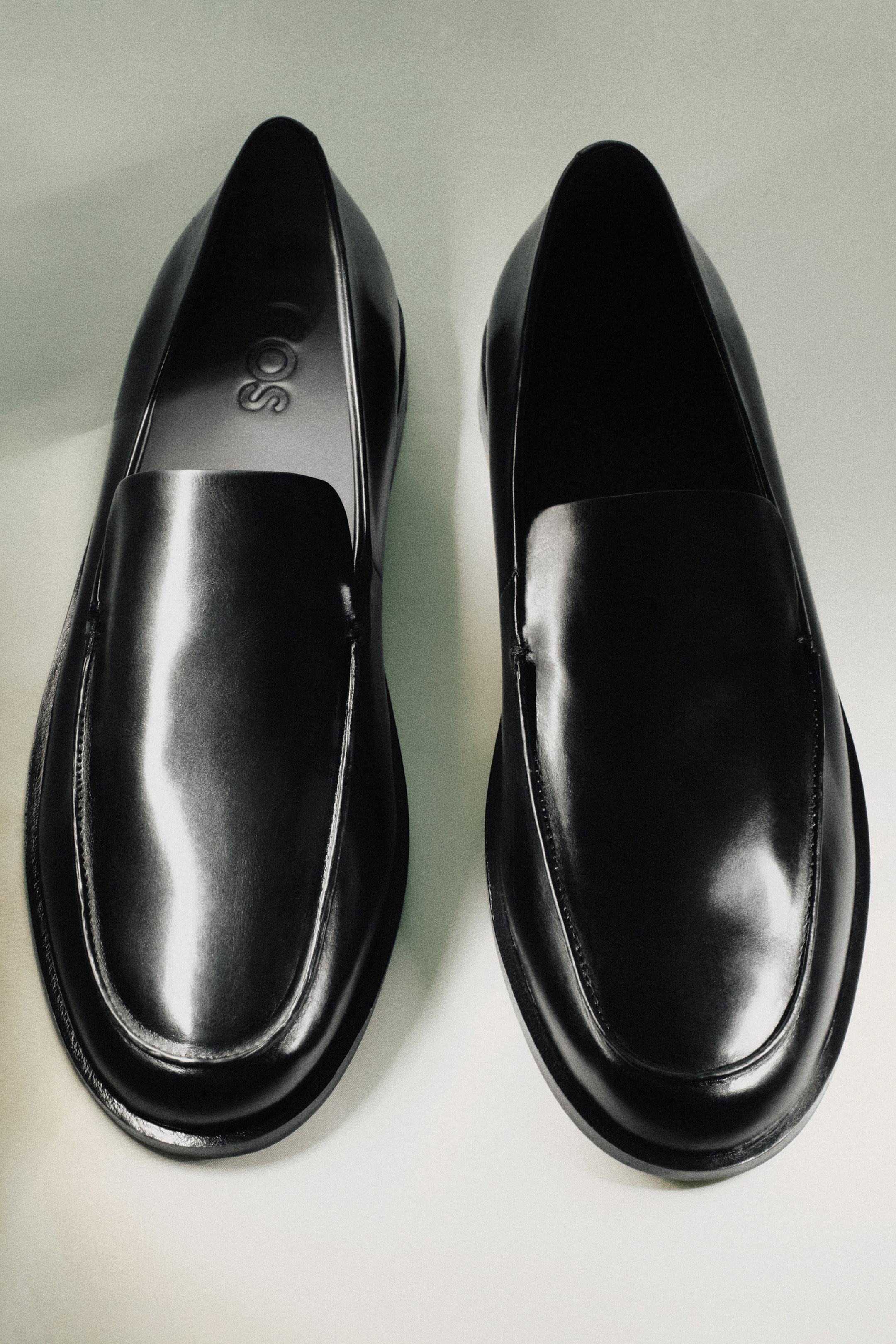 Visa större bild: Två svarta polerade läderloafers visas sida vid sida från en framifrån-ovanför vinkel, med rundade tår, synliga sömmar på tåns apron och "COS"-logotypen synlig på innersulan av den vänstra skon.