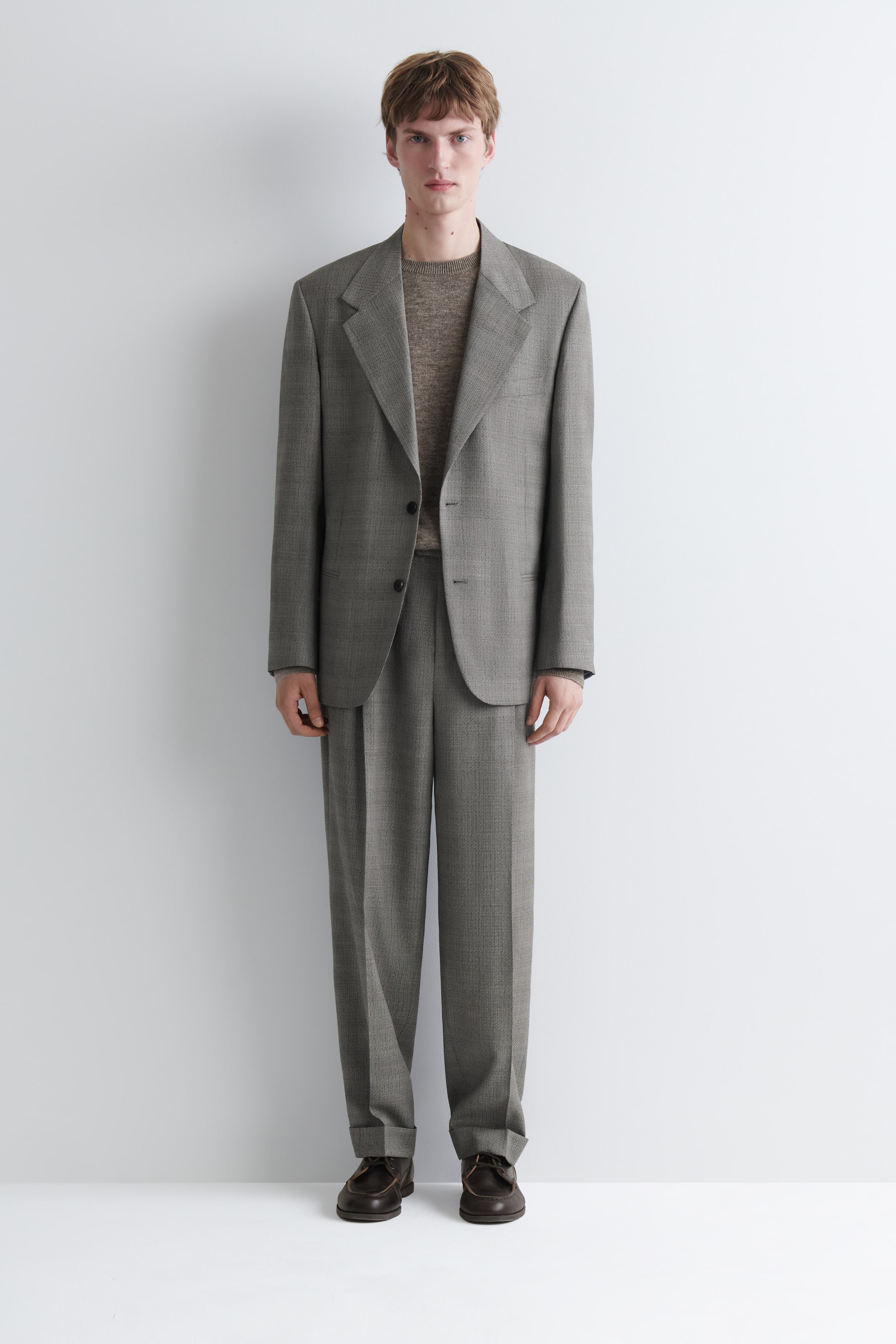 View larger image: The model is wearing a grey textured suit. The two-button jacket, featuring wide lapels, is layered over a brown crew neck sweater, while the pleated trousers are cuffed at the hem and paired with dark brown shoes.
