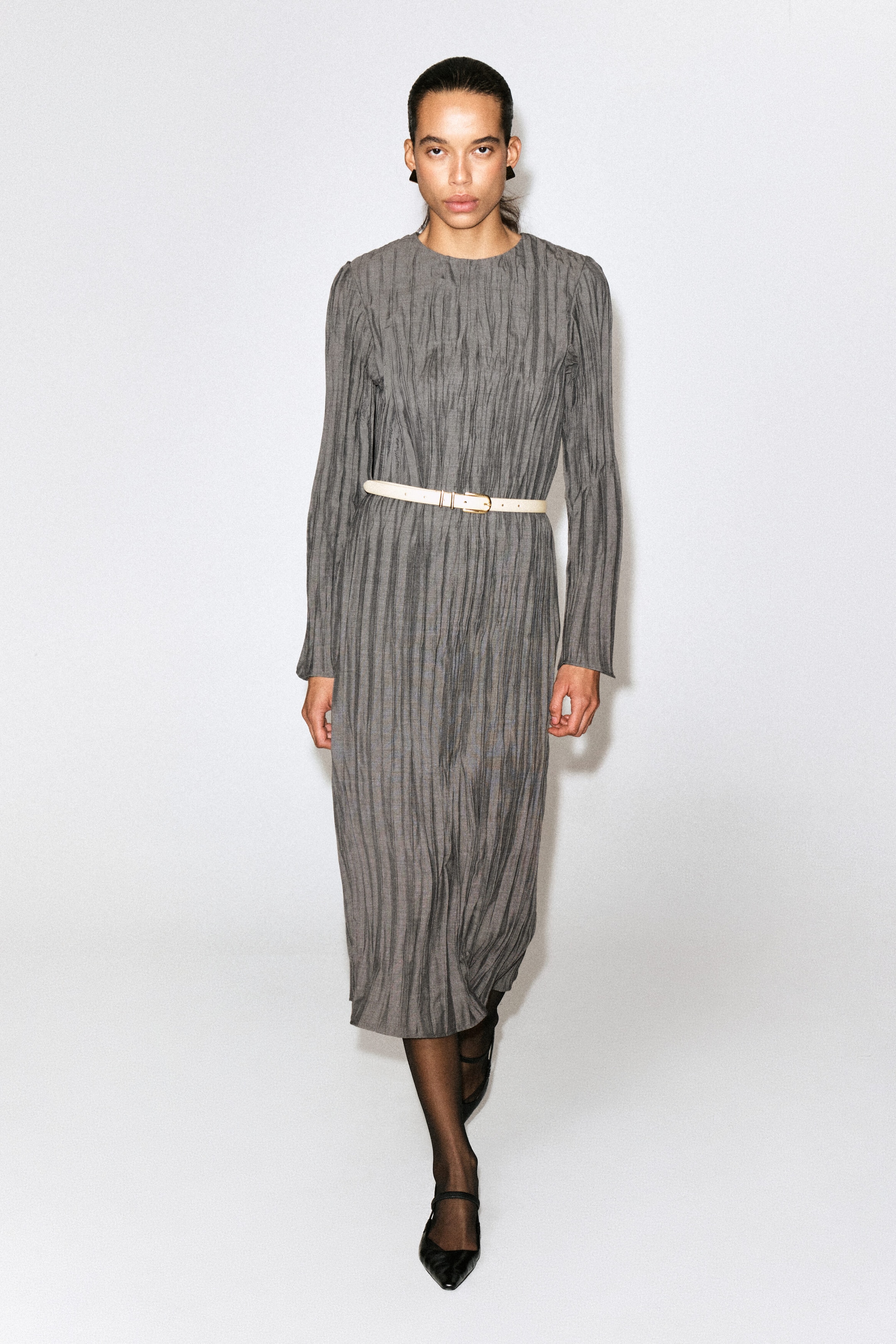 A gray midi dress featuring a vertical crinkled texture, a high crew neck, and long sleeves is shown from the front. The dress is styled with a thin cream belt that cinches the waist, defining its silhouette.