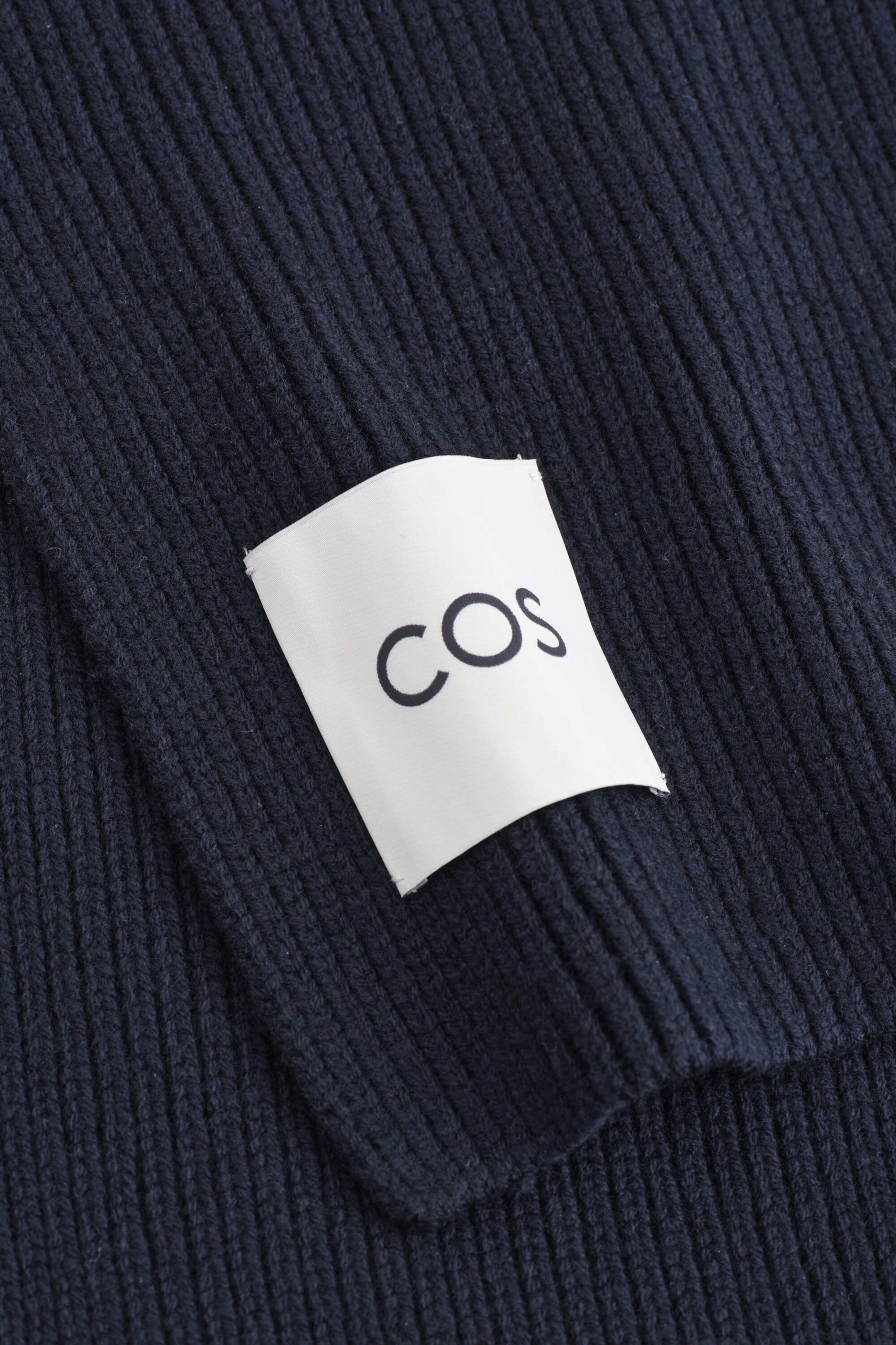 A navy blue ribbed knit item is displaying a white rectangular fabric label printed with the black brand name “COS”. The close-up shot highlights the texture of the vertical ribbing on the dark blue fabric. A navy blue ribbed knit item is displaying a white rectangular fabric label printed with the black brand name “COS”. The close-up shot highlights the texture of the vertical ribbing on the dark blue fabric.