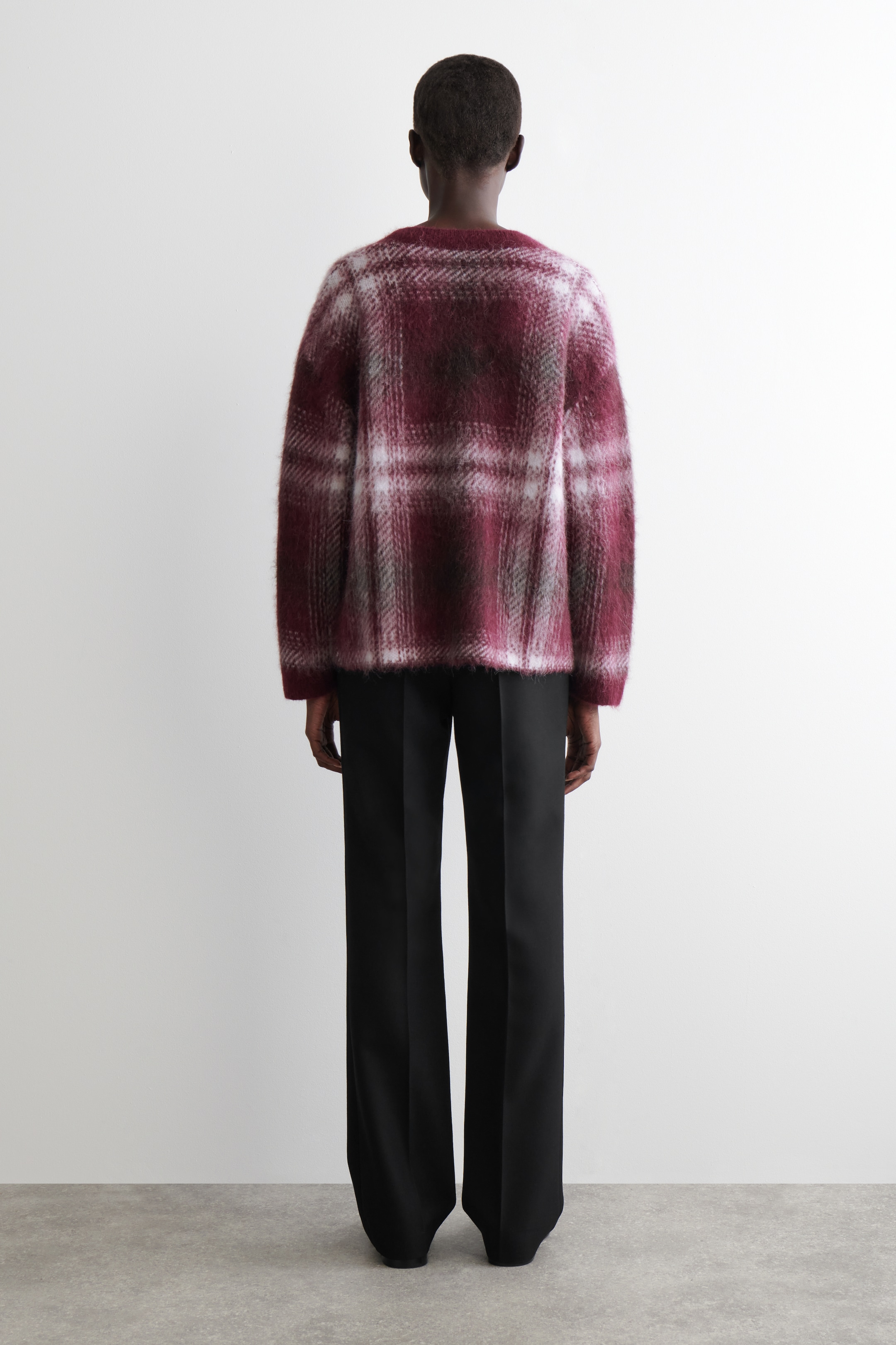 Grösseres Bild ansehen: Ein flauschiger, übergroßer Pullover in einem rot-weiß-grauen Karomuster wird von hinten präsentiert und verfügt über einen runden Halsausschnitt. Er wird mit locker sitzenden, schwarzen Hosen mit weitem Bein gestylt.