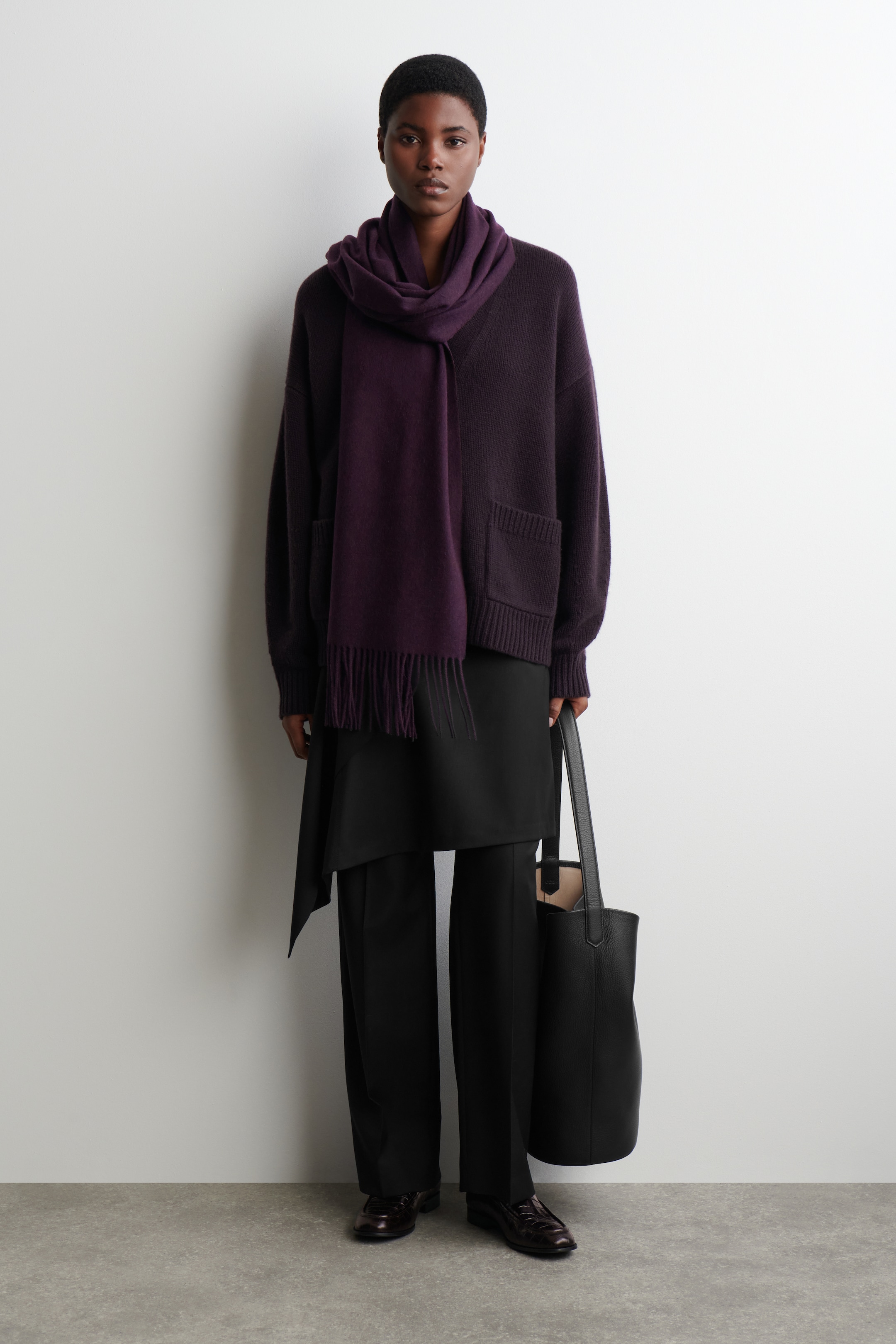 Agrandir l'image: Un cardigan surdimensionné violet foncé est porté, avec des poches plaquées à l'avant et de longues manches, superposé à un haut asymétrique noir. Une écharpe à franges violet foncé est drapée autour du cou, complétant un pantalon large noir, des mocassins marron foncé à motifs et un sac seau noir porté.
