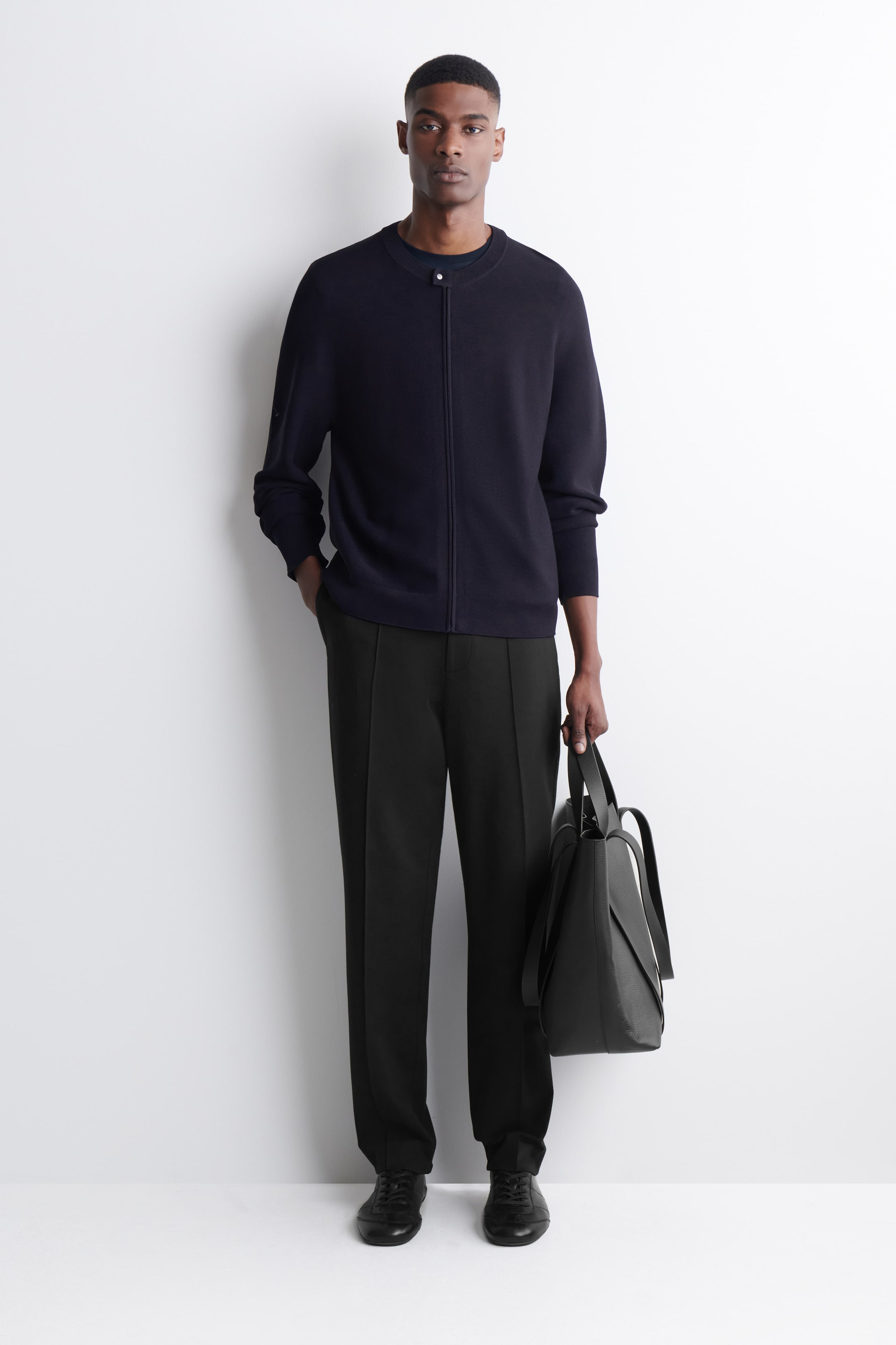 Преглед на по-голямо изображение: Представено е тъмносиньо плетено яке в стил бомбер, наслоено върху черна блуза с кръгло деколте и съчетано с черни прави панталони с изразителен преден шев. Комплектът е завършен с черни обувки с връзки и структурирана черна чанта тип "тоте", всички показани от фронтална перспектива.