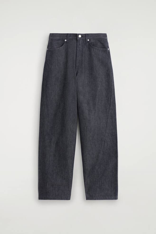 Donker indigo denim broek met wijde pijpen wordt van de VOORKANT gepresenteerd, met een ontspannen pasvorm en sculpturale overslagnaden die langs de buitenkant van de benen lopen. Een zilveren knoop is zichtbaar in de taille, samen met voorzakken en metalen klinknagels. - 1