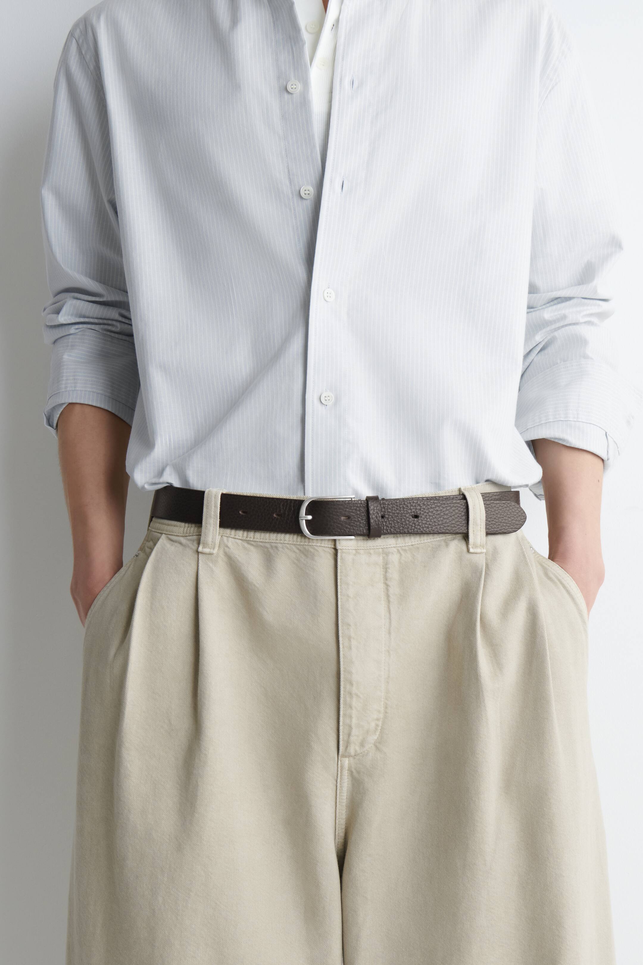 Grotere afbeelding bekijken: Een donkerbruine leren riem met een gestructureerde nerf en een gebogen, geborstelde zilverkleurige gesp wordt door het model gedragen. De riem houdt een beige plooi broek op zijn plaats, met een licht gestreept hemd ingestopt in de tailleband.