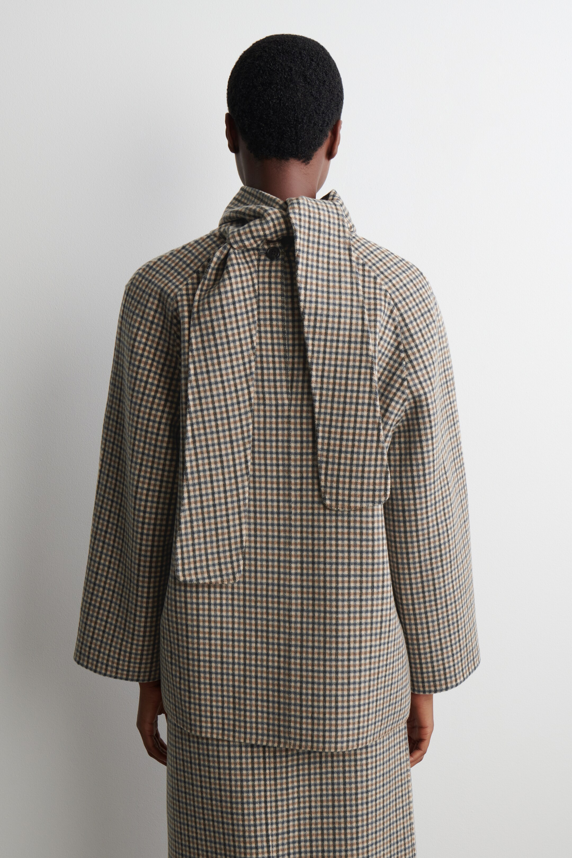 Ver imagen más grande: Una chaqueta corta en un patrón de cuadros pequeños en tonos marrones, beige y oscuros se presenta desde la parte trasera, con un cuello alto con cierre de lazo largo anudado en la nuca y mangas anchas y rectas. Una falda a juego también se lleva debajo de la chaqueta.