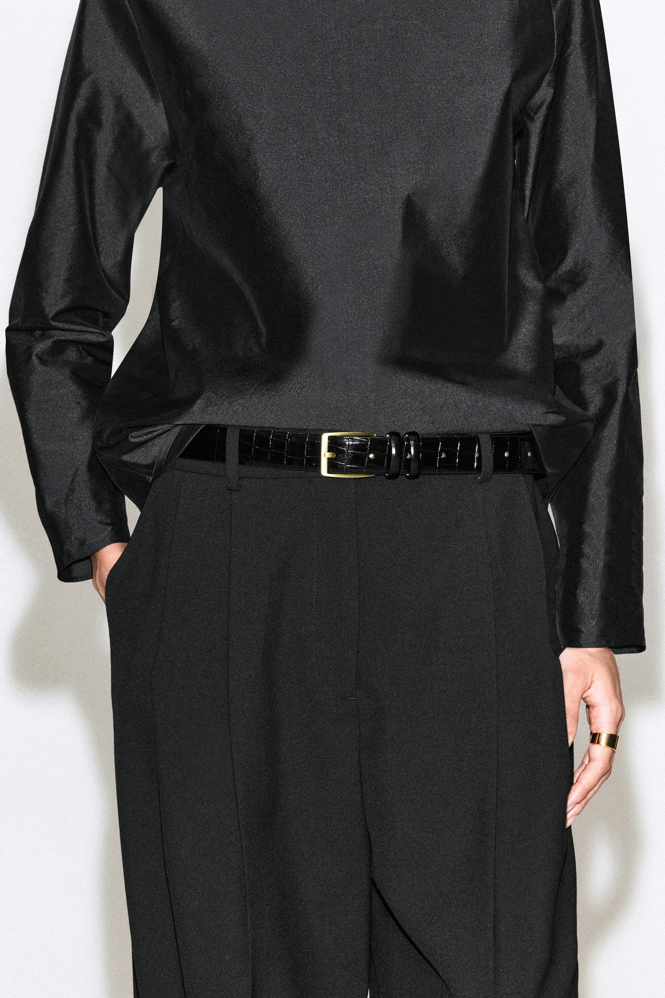 A black croc-effect leather belt featuring a gold-tone buckle is being worn by the model. The belt is threaded through the loops of black trousers and styled with a tucked-in black satin top.