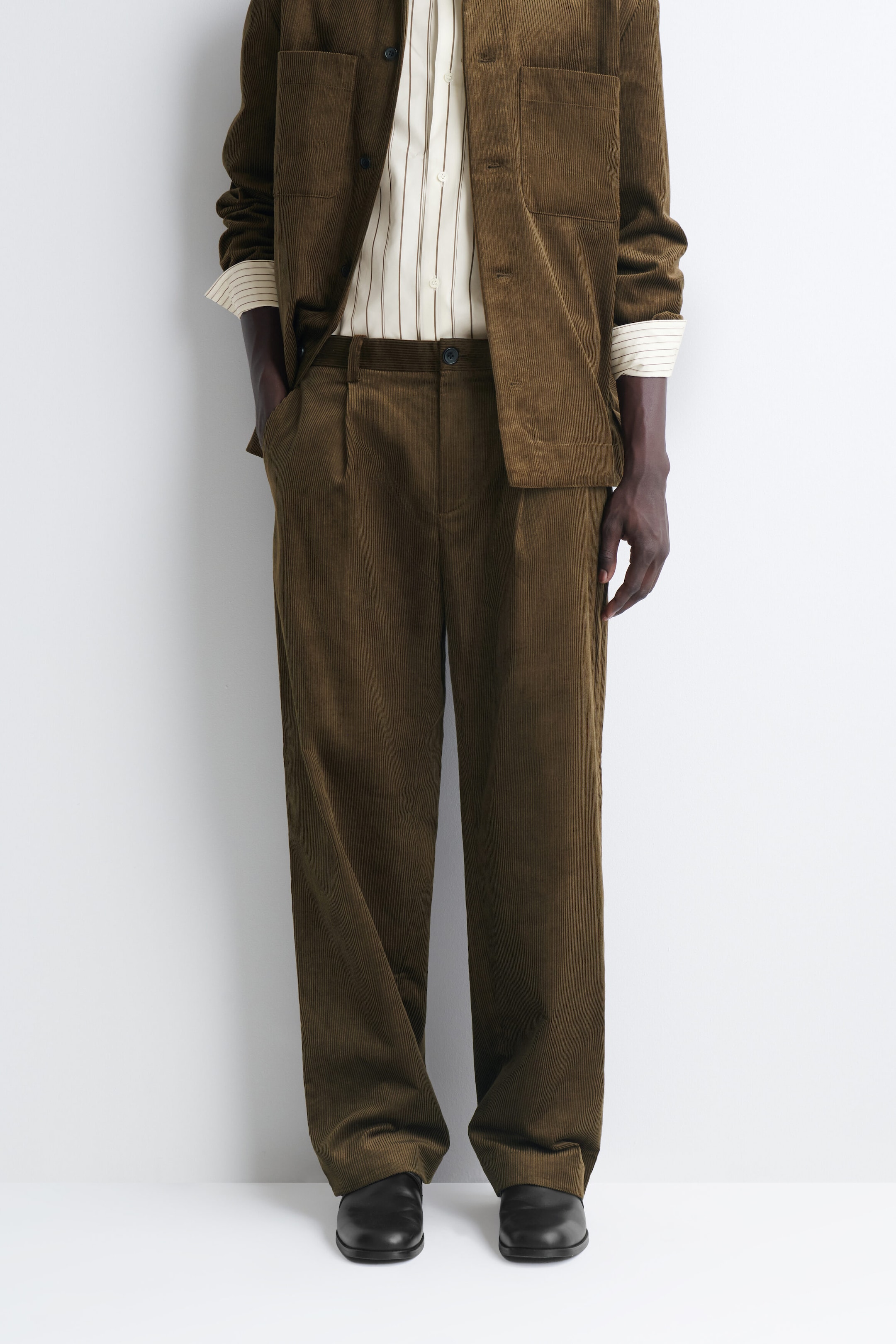 The Pleated Relaxed-Fit Tapered Corduroy Pants are shown from the front, featuring rich olive-brown corduroy fabric with front pleats, belt loops, and a relaxed, tapered silhouette. They are styled with a tucked-in striped shirt, a matching open corduroy overshirt, and black leather shoes. The Pleated Relaxed-Fit Tapered Corduroy Pants are shown from the front, featuring rich olive-brown corduroy fabric with front pleats, belt loops, and a relaxed, tapered silhouette. They are styled with a tucked-in striped shirt, a matching open corduroy overshirt, and black leather shoes.