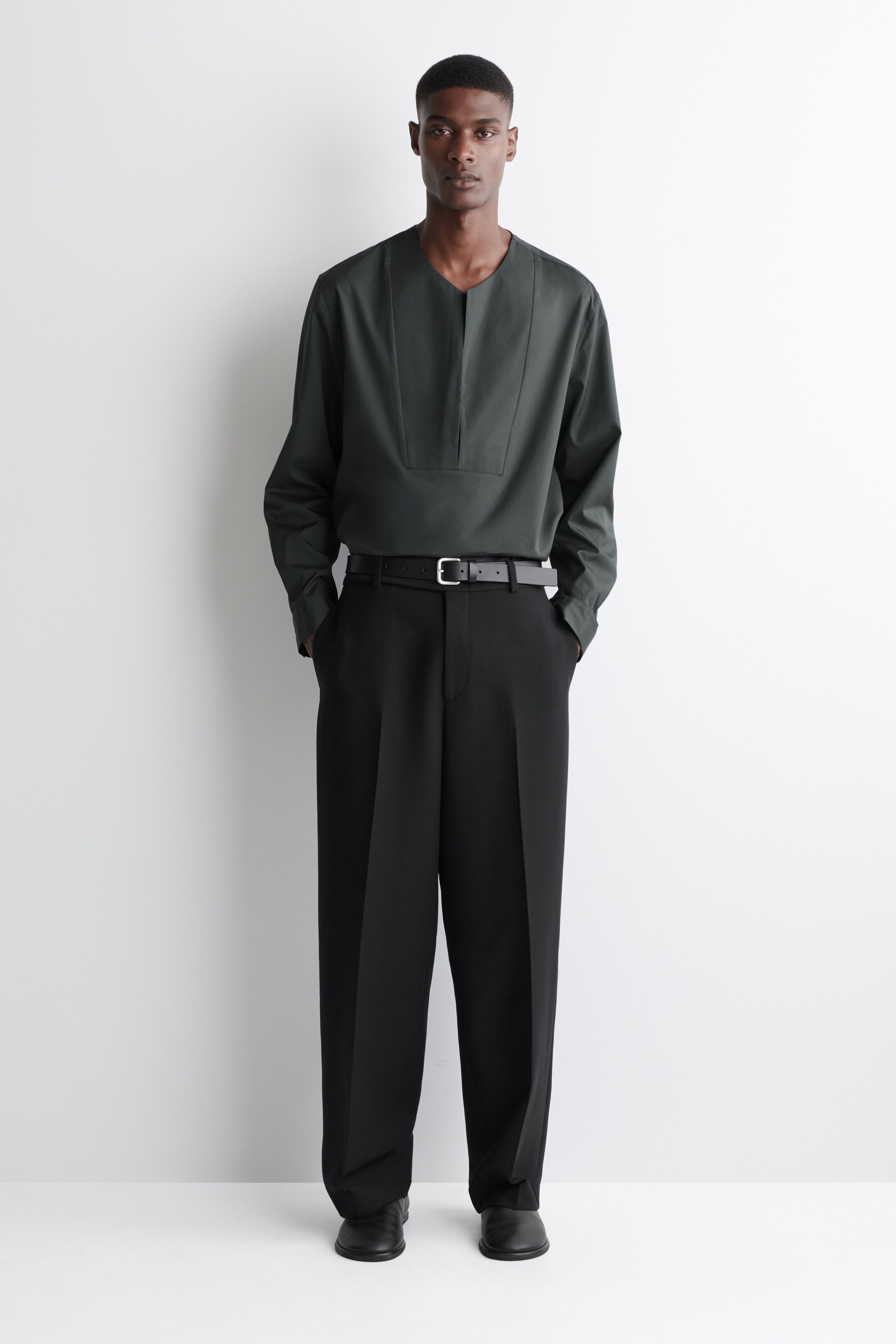 Ver imagen más grande: Una camisa de manga larga verde oscuro con cuello en V y un panel en el pecho se lleva con unos pantalones negros de pierna ancha. Los pantalones están ceñidos en la cintura por un cinturón negro y cuentan con pliegues frontales prominentes y un ajuste holgado, todo presentado hacia adelante.
