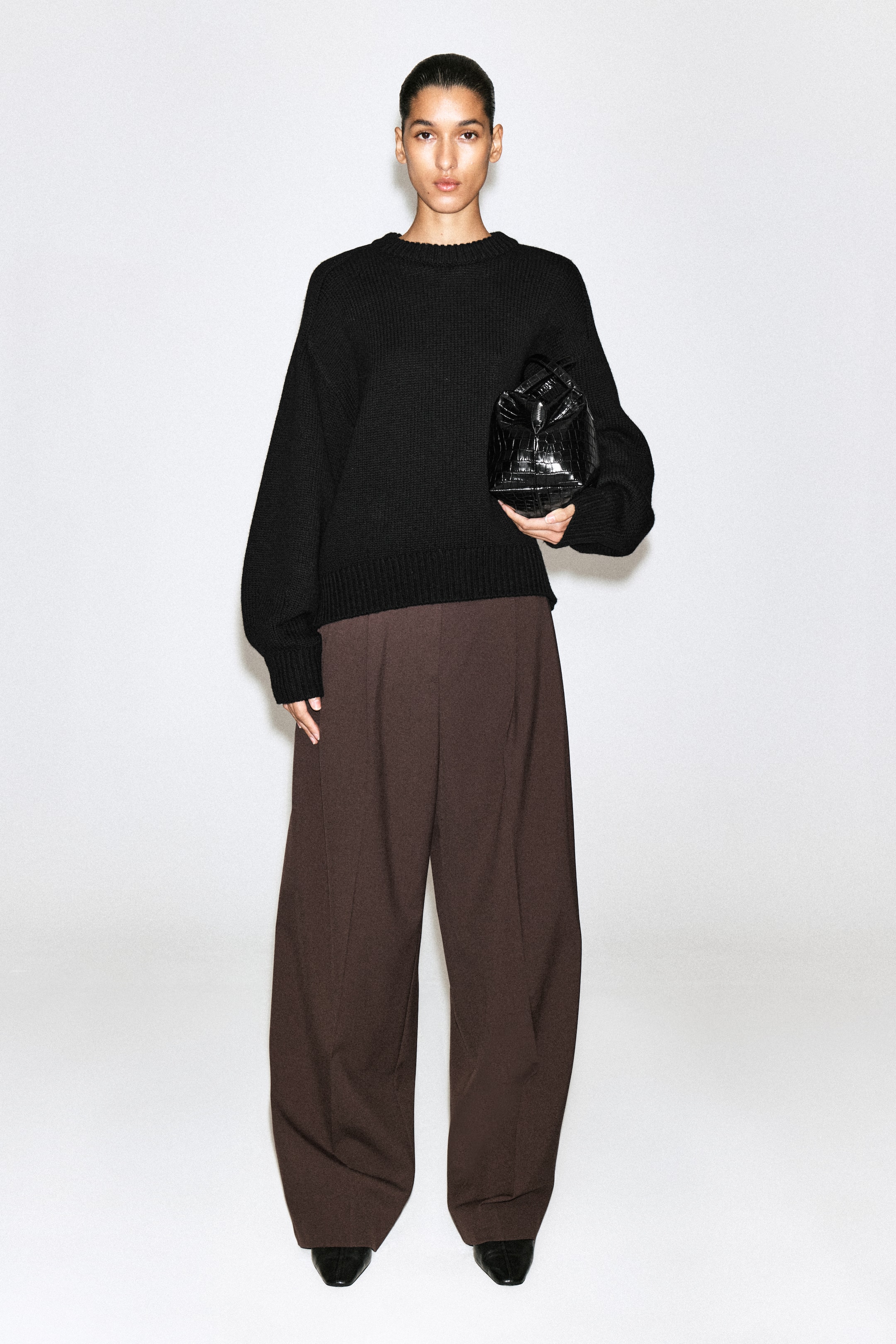 Visualizza l'immagine ingrandita: Un maglione nero in maglia con girocollo, polsini e orlo a coste, abbinato a pantaloni ampi marrone scuro in una vista frontale. Il maglione dalla vestibilità rilassata è completato da una borsa strutturata nera con finitura effetto coccodrillo, creando un look coordinato e minimalista.