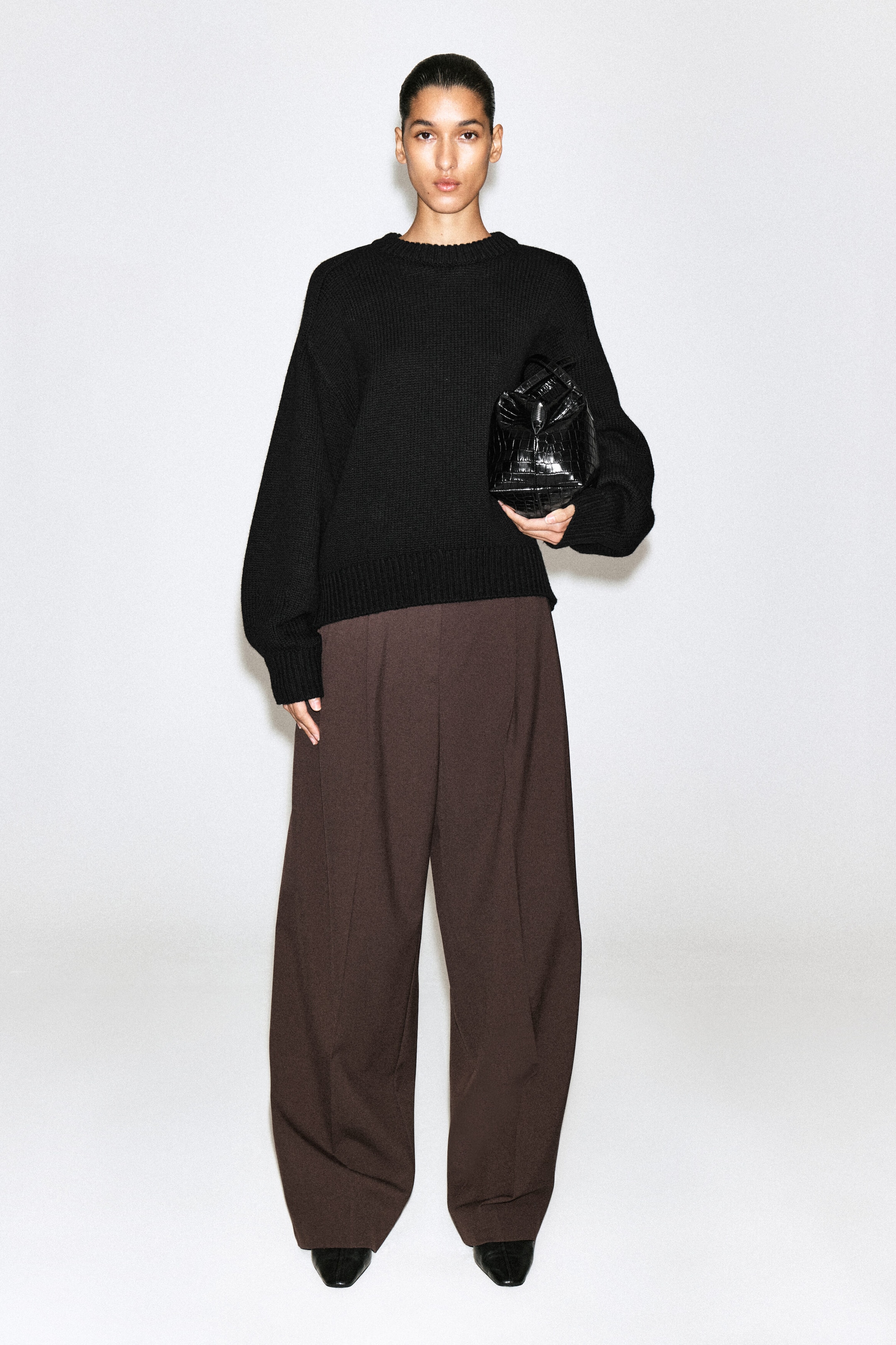 A black crew-neck knitted sweater featuring ribbed cuffs and hem is paired with dark brown wide-leg trousers in a front-facing view. The relaxed-fit sweater is styled with a structured black crocodile-embossed handbag, creating a coordinated, minimalist look.