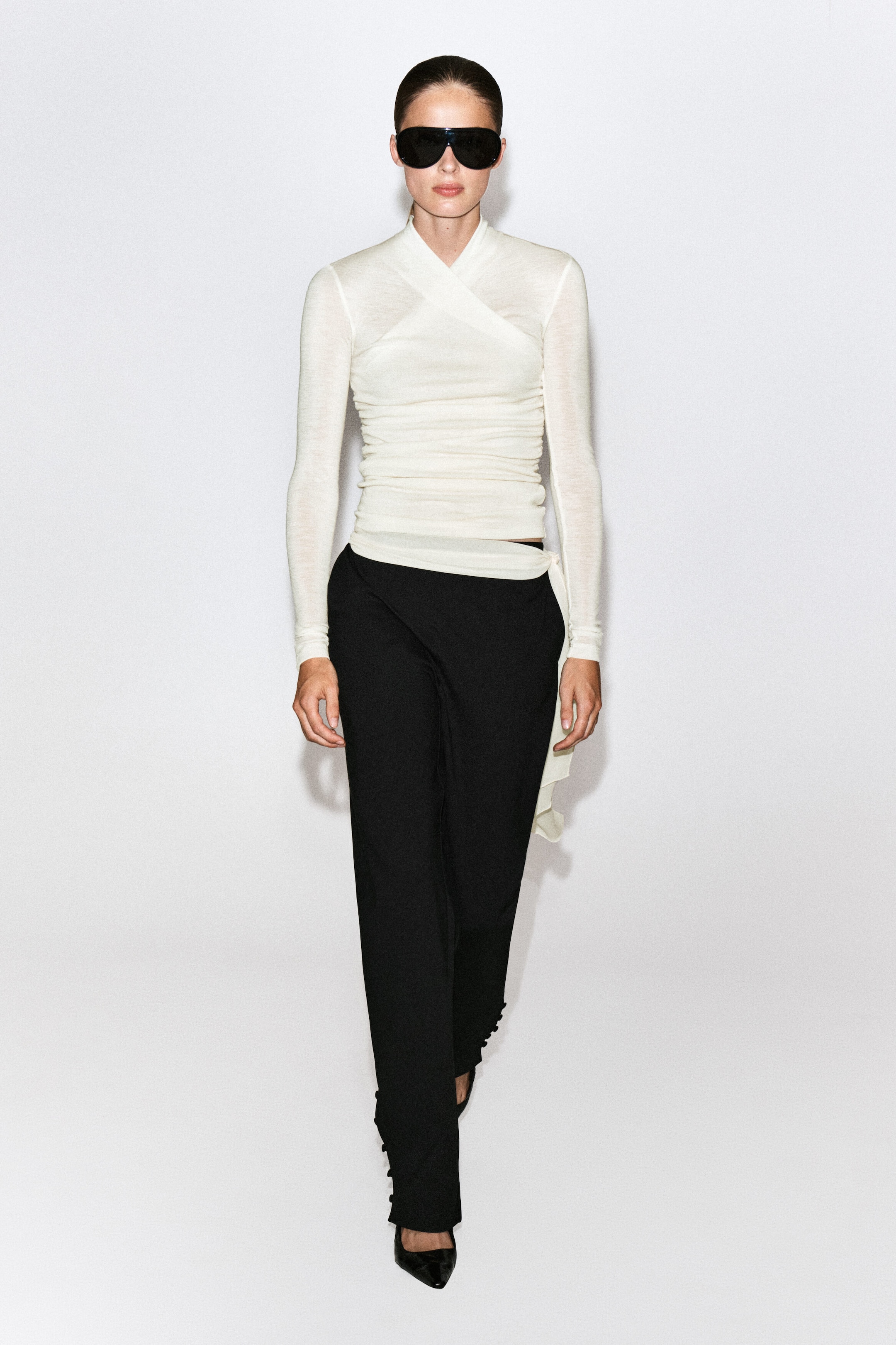 An off-white, long-sleeved wrap top featuring a crossed shawl collar and gathered detailing along the bodice is being worn by the model. The slim-fit garment is styled with black trousers. An off-white, long-sleeved wrap top featuring a crossed shawl collar and gathered detailing along the bodice is being worn by the model. The slim-fit garment is styled with black trousers.