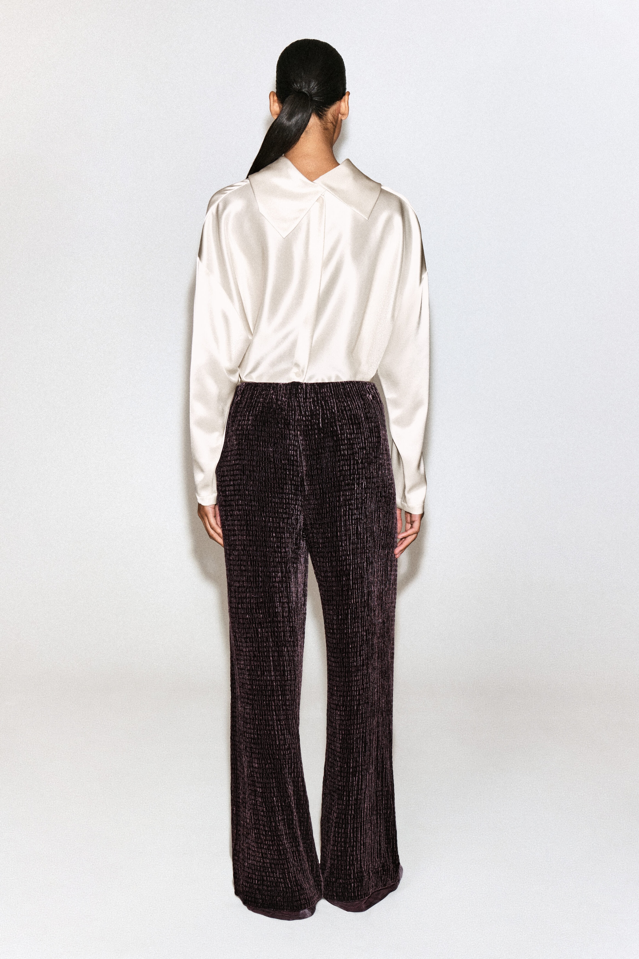 A cream-colored satin blouse and dark plum textured trousers are shown from the back, highlighting their fit and design. The long-sleeve blouse features a draped collar detail and is tucked into the wide-leg, crinkled velvet trousers. A cream-colored satin blouse and dark plum textured trousers are shown from the back, highlighting their fit and design. The long-sleeve blouse features a draped collar detail and is tucked into the wide-leg, crinkled velvet trousers.