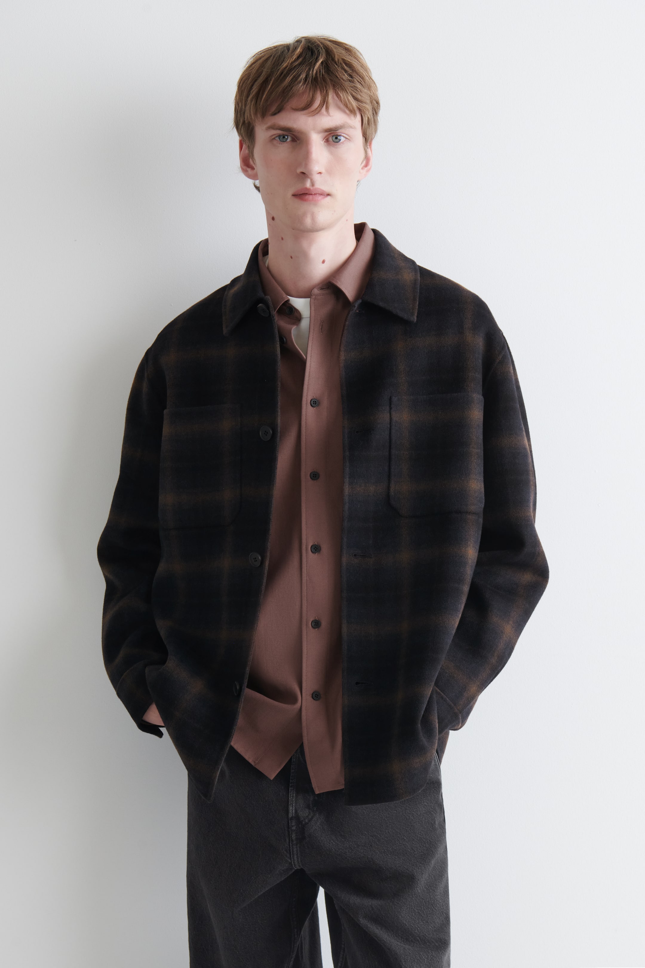Grotere afbeelding bekijken: Een donkerbruine geruite overshirt, met een puntkraag, knoopsluiting en twee opgestikte borstzakken, wordt gedragen door het model. Het is gedragen over een effen bruine shirt, terwijl de handen van het model in de zakken van het overshirt rusten.