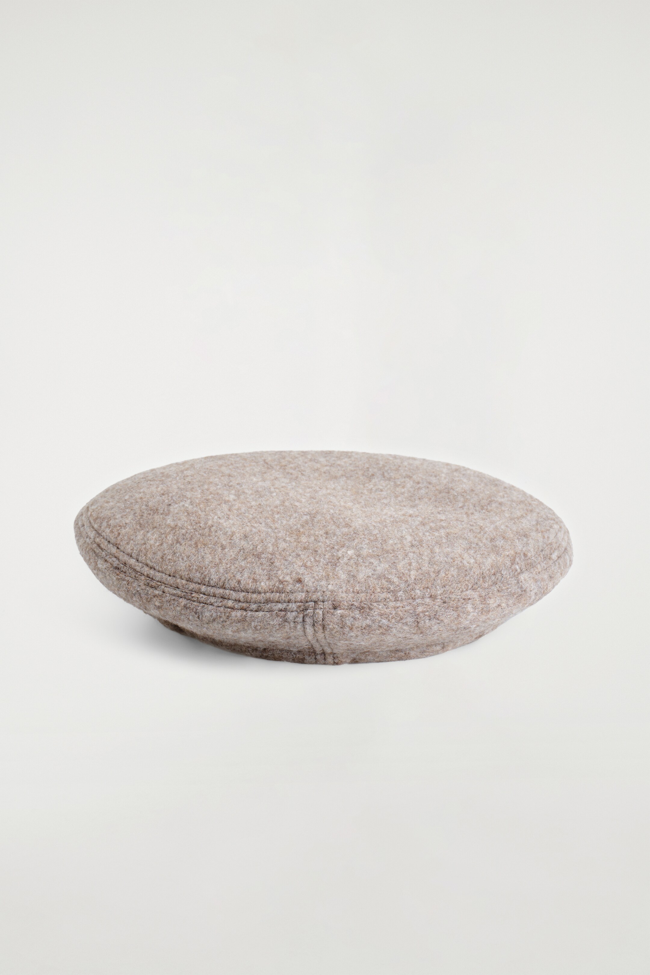 Agrandir l'image: Un béret beige-mélange est posé à plat, exhibant son tissu texturé et les lignes subtiles de couture le long de son bord arrondi.