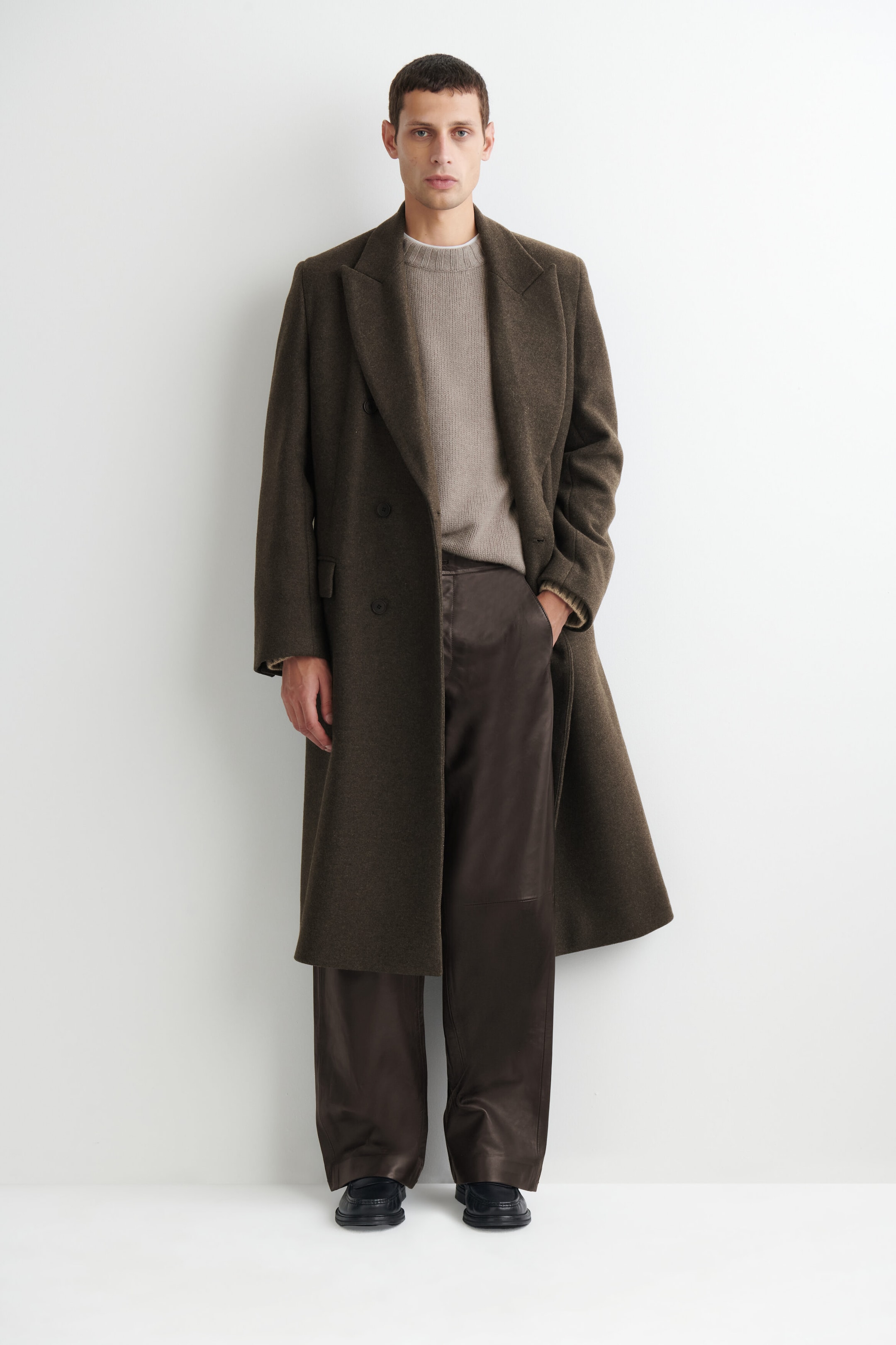 These dark brown leather relaxed-fit straight-leg pants drape in a loose, straight silhouette from the front, pooling slightly over black loafers. The pants feature a smooth leather finish, with one hand tucked into a side pocket, styled alongside a partially tucked grey sweater and an open overcoat. These dark brown leather relaxed-fit straight-leg pants drape in a loose, straight silhouette from the front, pooling slightly over black loafers. The pants feature a smooth leather finish, with one hand tucked into a side pocket, styled alongside a partially tucked grey sweater and an open overcoat.
