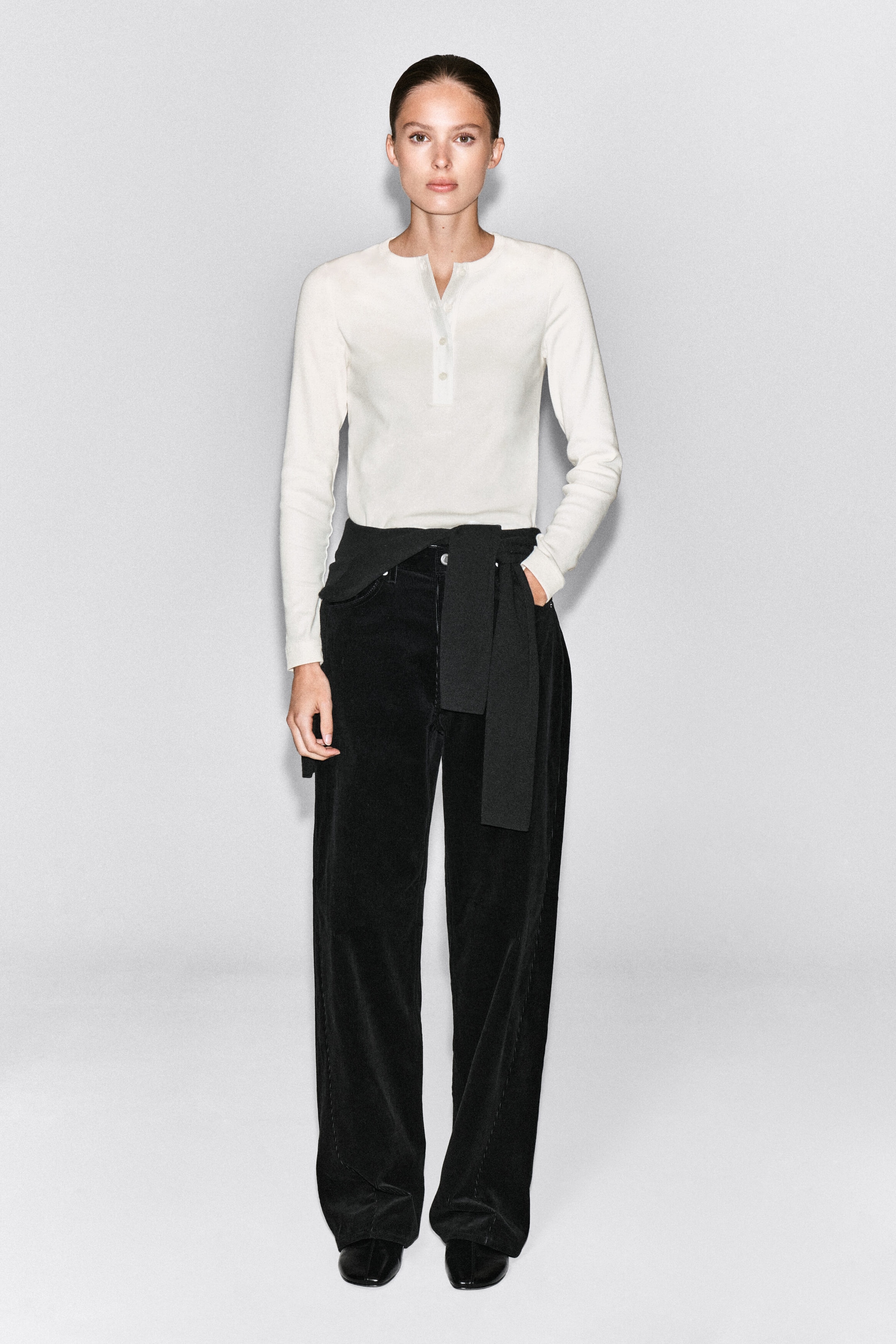 An off-white ribbed cotton henley top is being worn by the model, featuring long sleeves and a buttoned placket at the neckline. The top is styled tucked into wide-leg black trousers with a black garment tied around the waist. An off-white ribbed cotton henley top is being worn by the model, featuring long sleeves and a buttoned placket at the neckline. The top is styled tucked into wide-leg black trousers with a black garment tied around the waist.