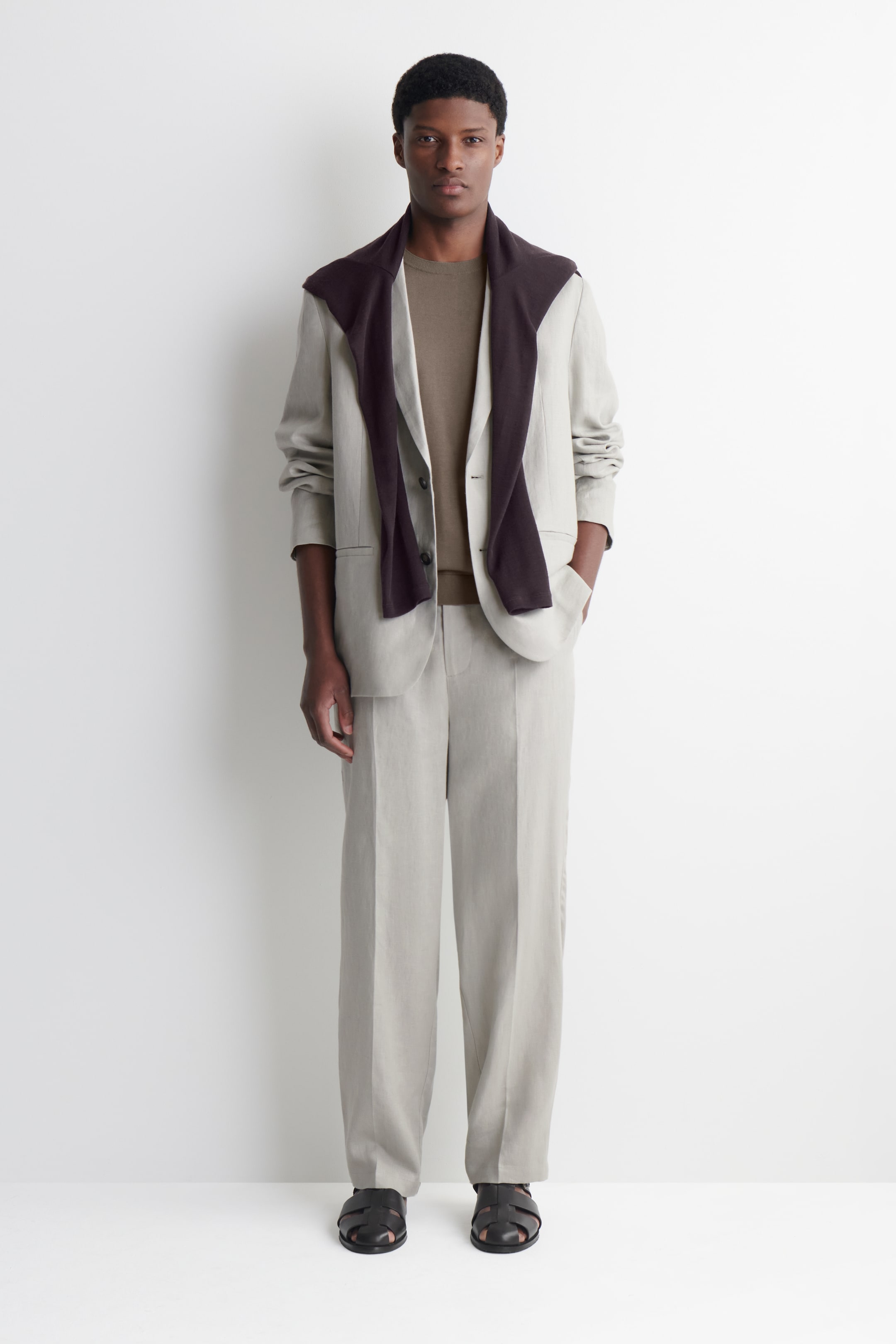 View larger image: A light beige single-breasted blazer, featuring long sleeves and notch lapels, is styled open over a brown crew-neck top. Coordinating wide-leg trousers complement the blazer, while a dark brown knit is elegantly draped over the blazer's shoulders. Black caged sandals complete the ensemble.