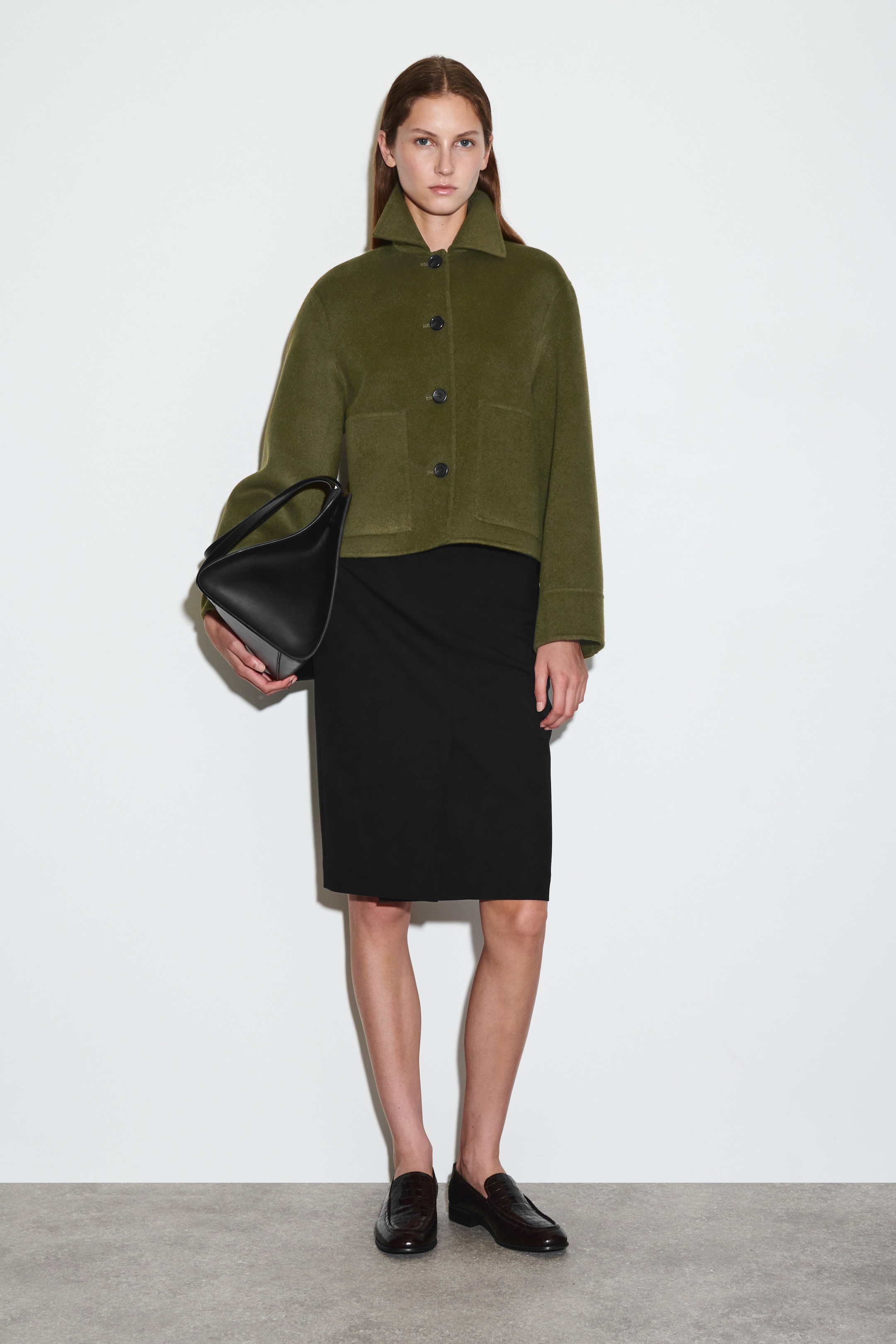 Agrandir l'image: Une veste courte vert olive est présentée de face, avec une coupe ample et carrée, un col pointu, trois boutons foncés à l'avant et deux grandes poches plaquées à l'avant. Elle est portée par-dessus une jupe noire longueur genou et est assortie d'un sac à main structuré noir tenu à la main.