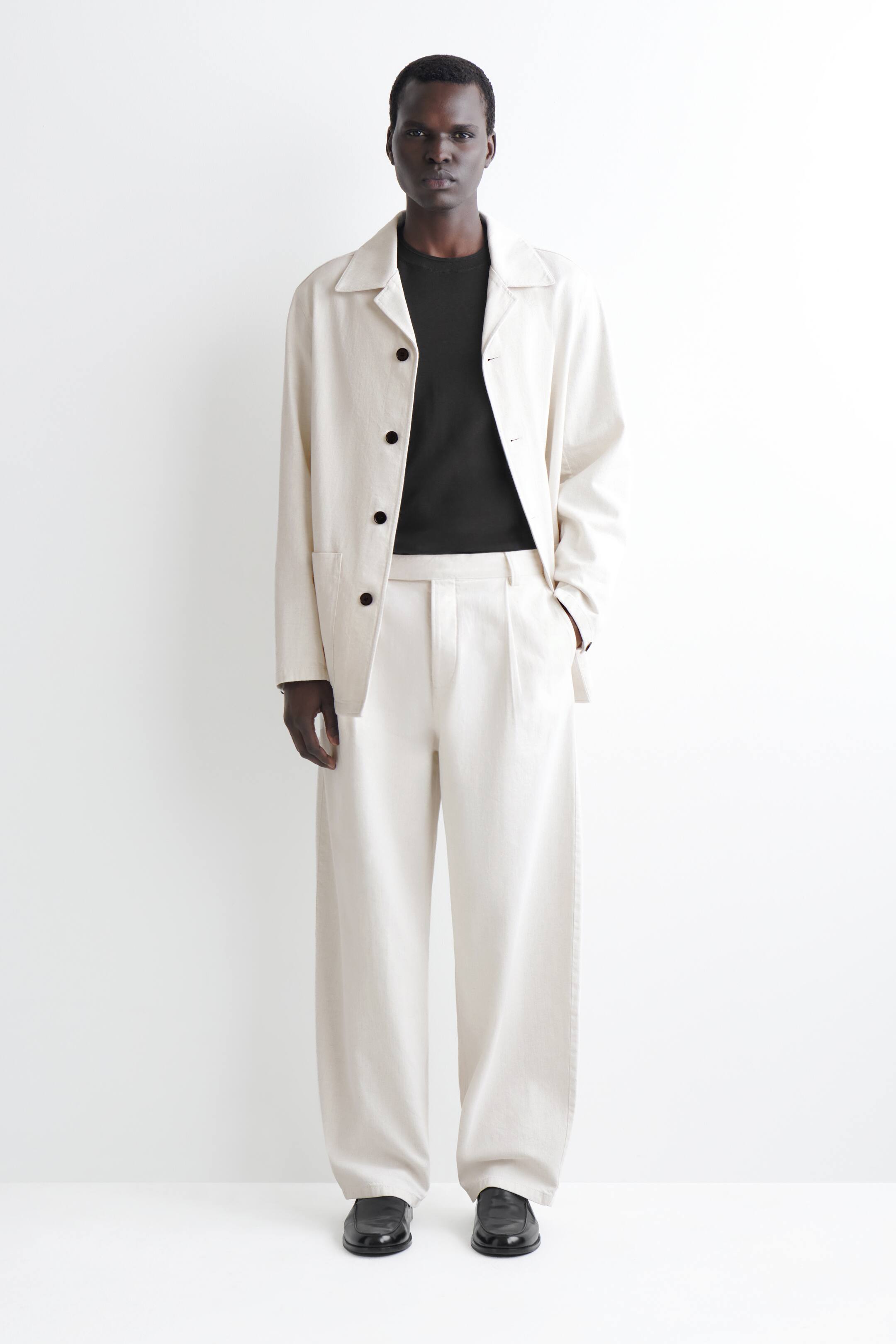 הצגת תמונה גדולה יותר: ג'קט עבודה בצבע קרם בהיר נלבש פתוח מעל חולצת טי שירט שחורה עם צווארון עגול, כולל צווארון פתוח, ארבעה כפתורים כהים ושני כיסי טלאי קדמיים. מכנסיים תואמים בצבע קרם בהיר, רחבים עם קפלים קדמיים משלימים את ההופעה, מעוצבים עם נעלי לופרס שחורות.