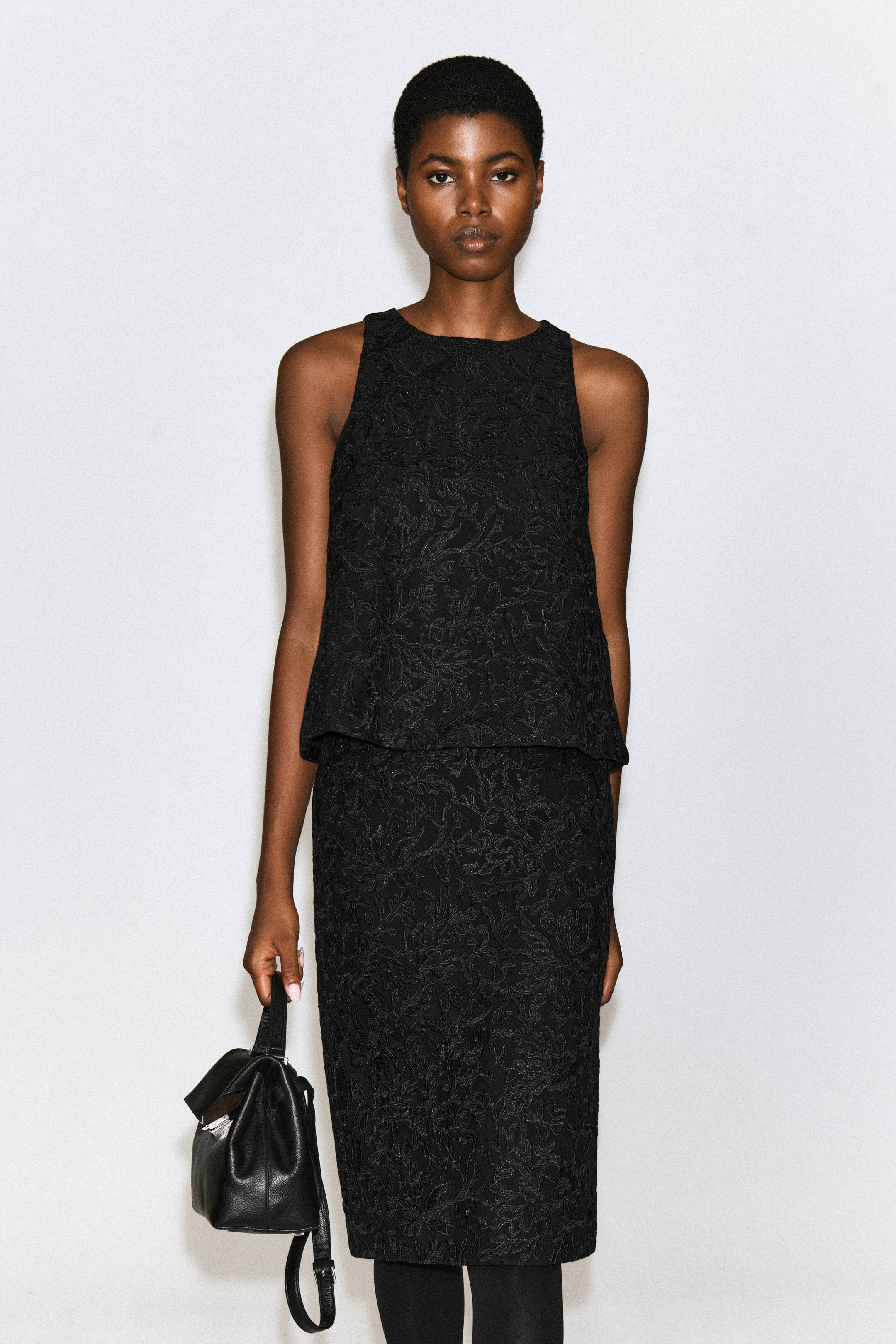 View larger image: A sleeveless black top featuring textured floral embroidery is being worn by the model, paired with a matching lace skirt. The model is also holding a black leather handbag.