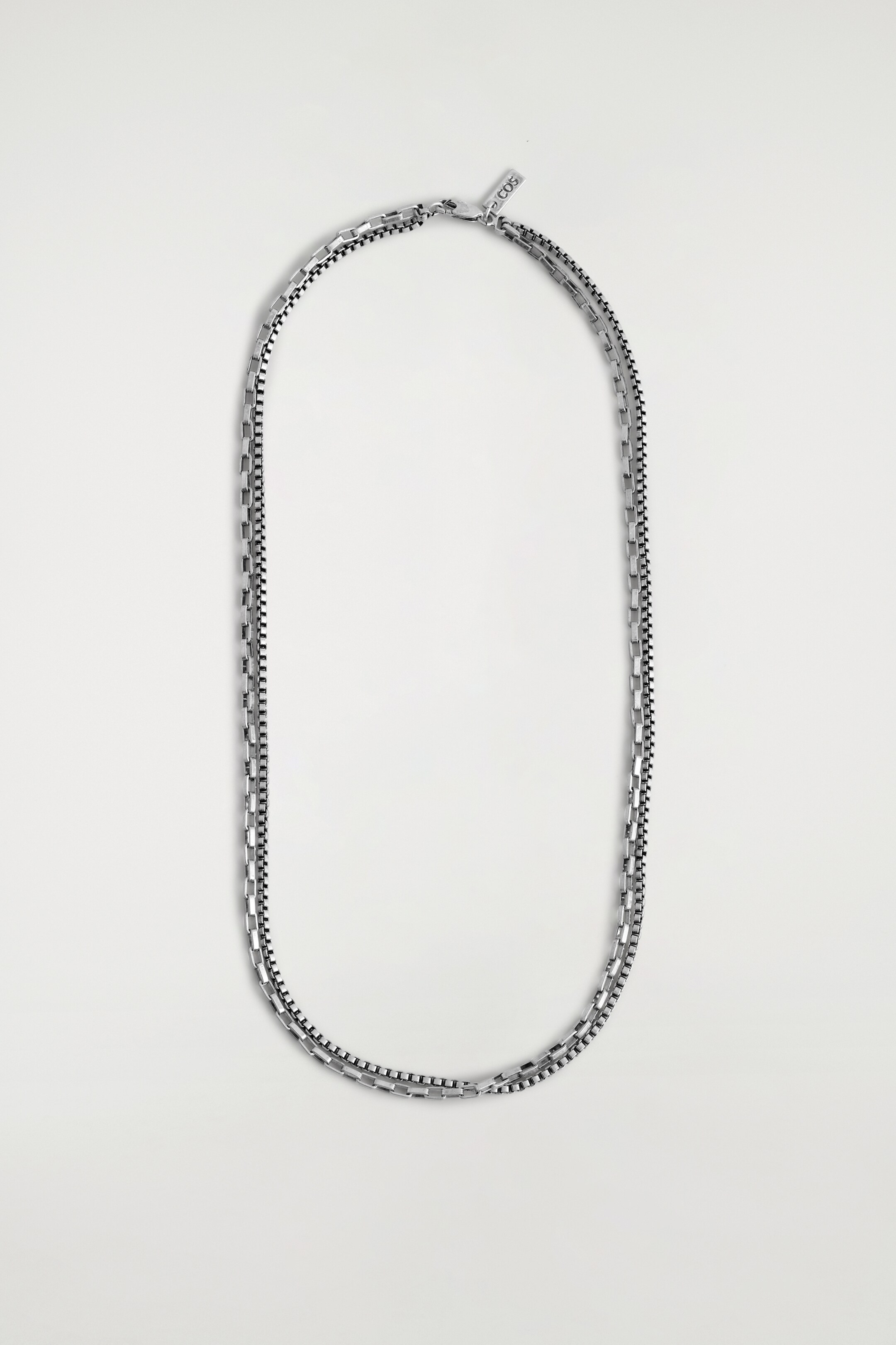 Visualizza immagine più grande: Una collana placcata in argento è esposta frontalmente, caratterizzata da due catene a maglia quadrata distinte e sovrapposte, collegate in alto da un unico moschettone e un piccolo pendente rettangolare.
