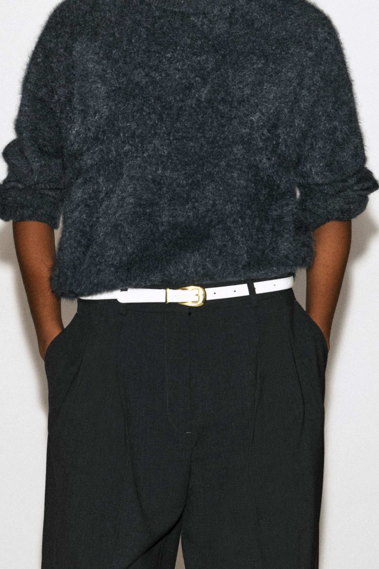 A white leather belt with a sculpted gold-tone buckle is being worn by the model, threaded through the belt loops of black trousers. The belt is styled with a dark grey fuzzy knit sweater tucked into the waist. - 1