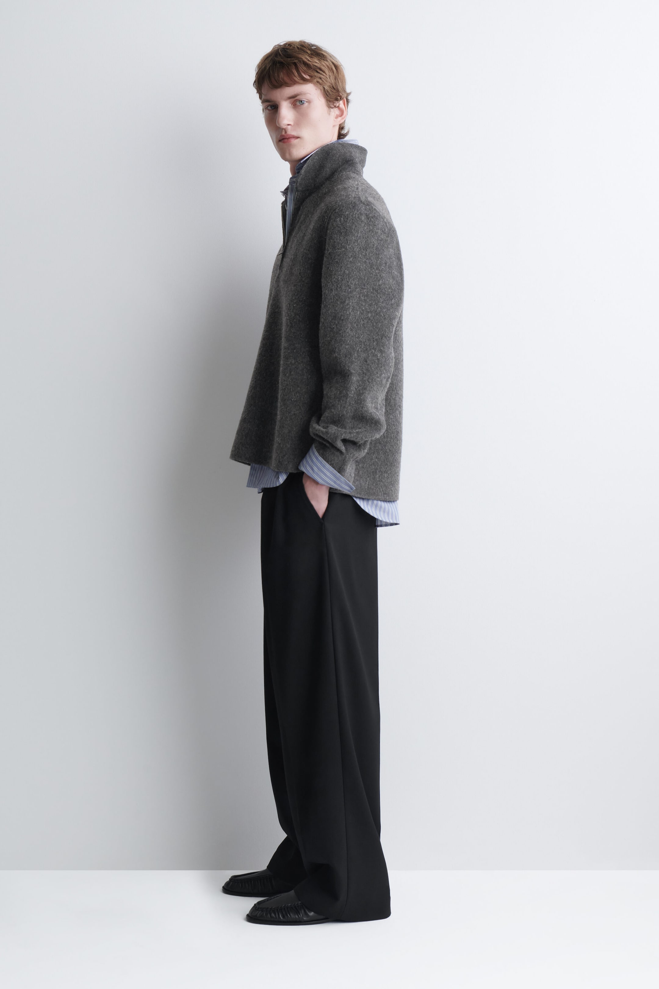 A gray, textured high-neck knit sweater is being worn by the model, layered over a light blue striped shirt. The sweater is paired with black wide-leg trousers, with the model standing in profile.