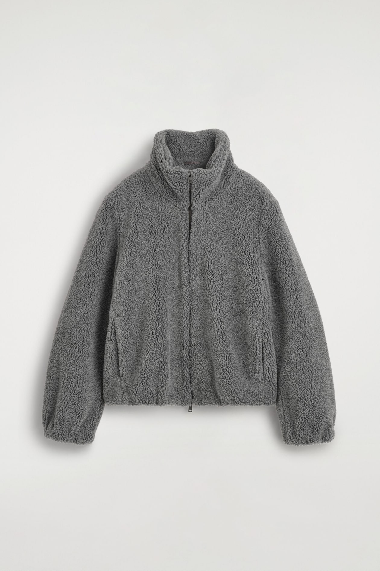 A grey teddy fleece jacket is shown from the front, highlighting a highly textured fleece material, a high funnel neck, and a centre front zipper. The jacket features long sleeves with gathered cuffs and side welt pockets.