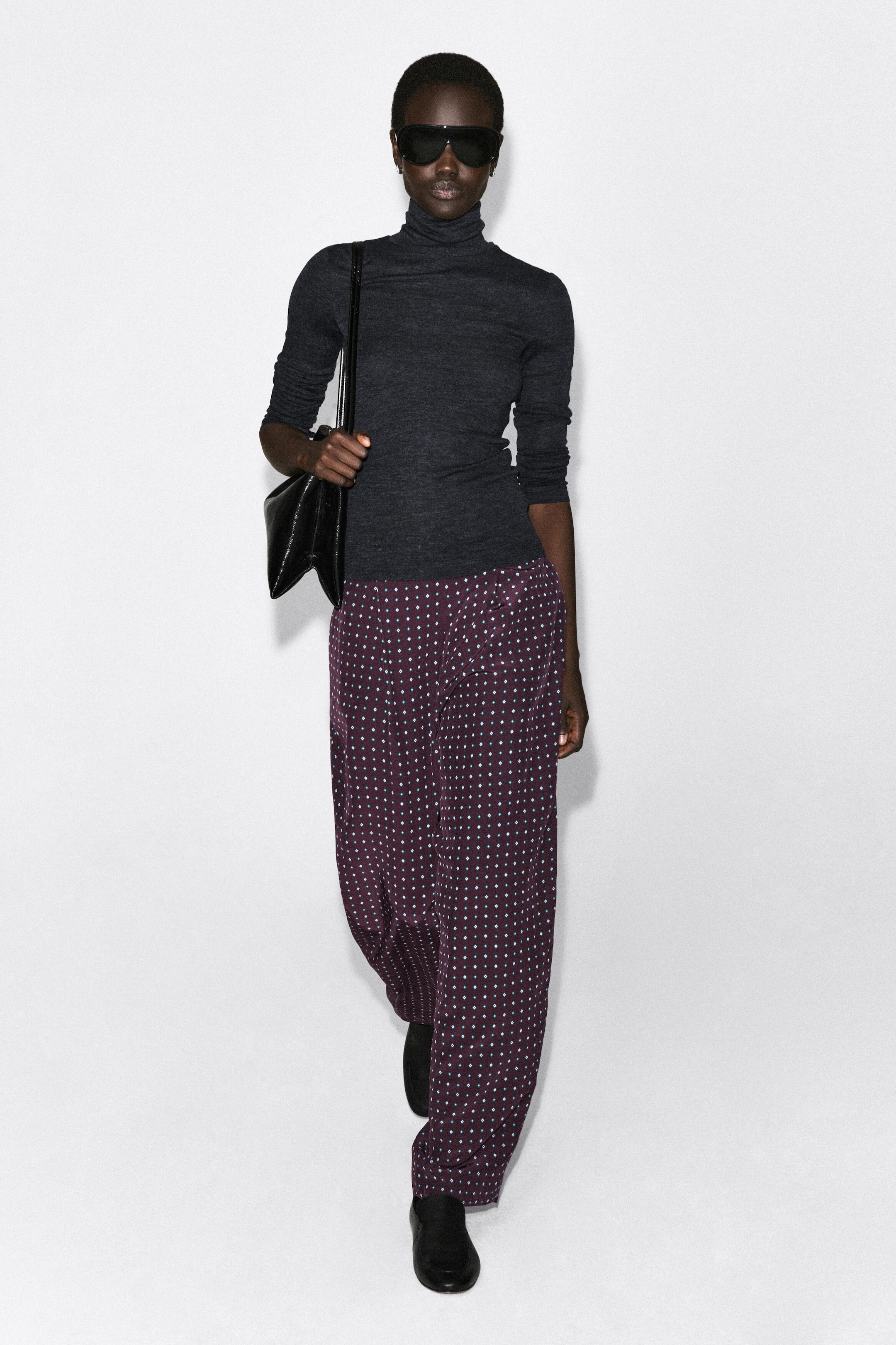 Ce pantalon droit à imprimé losanges est présenté de face, mettant en valeur une coupe ample et fluide dans un tissu prune profond orné d’un motif répété de petits losanges bleu sarcelle. Le pantalon tombe librement jusqu’à la cheville, soulignant sa silhouette droite, et est associé à un col roulé gris foncé.